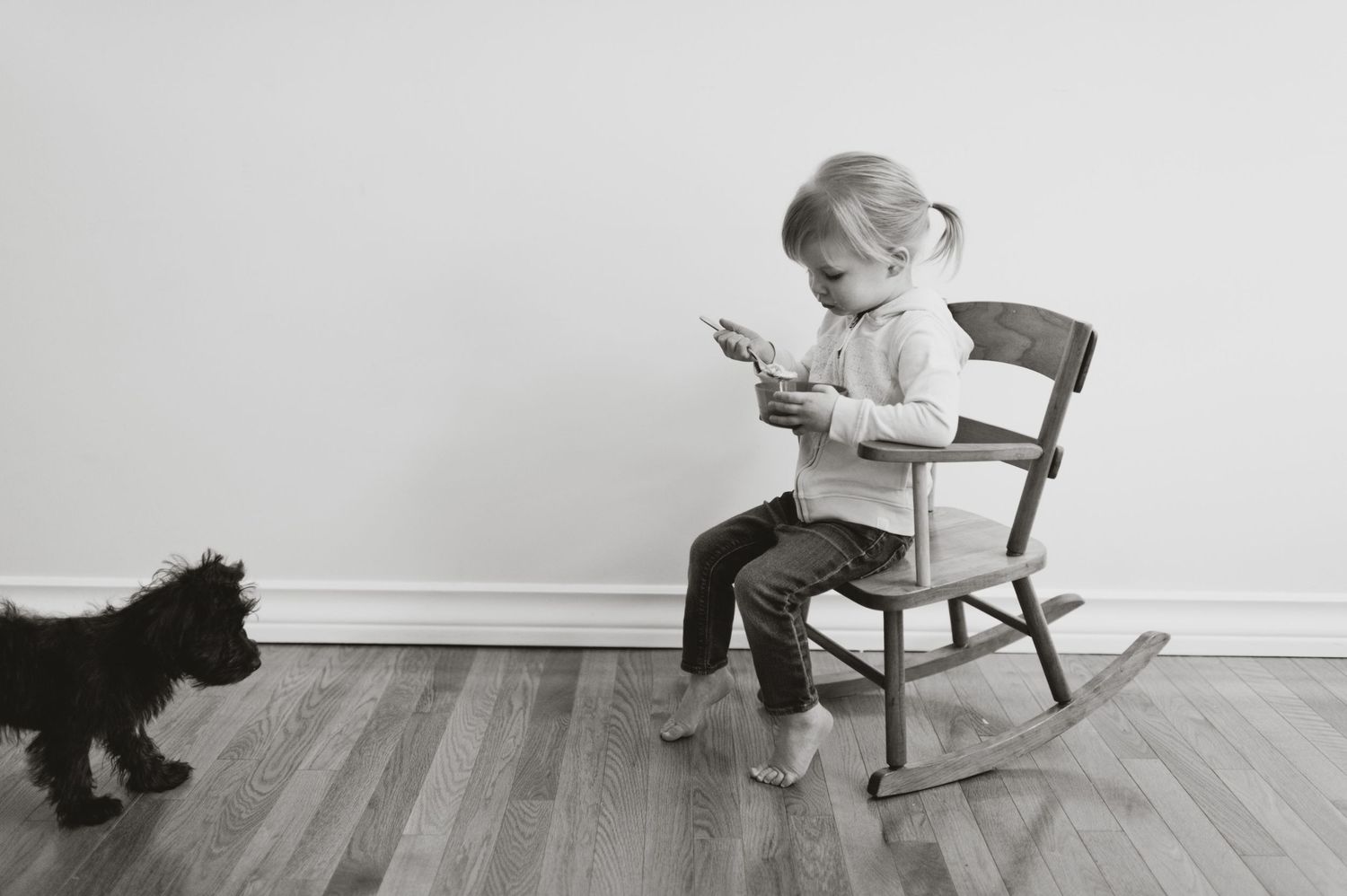 Toddler sitting barefoot in a wooden rocking chair, focused on an object in hand, while a small dog looks on. Minimalist black and white