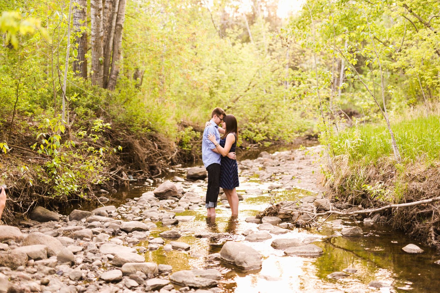 Couple kissing barefoot in a rocky creek surrounded by bright spring greenery.