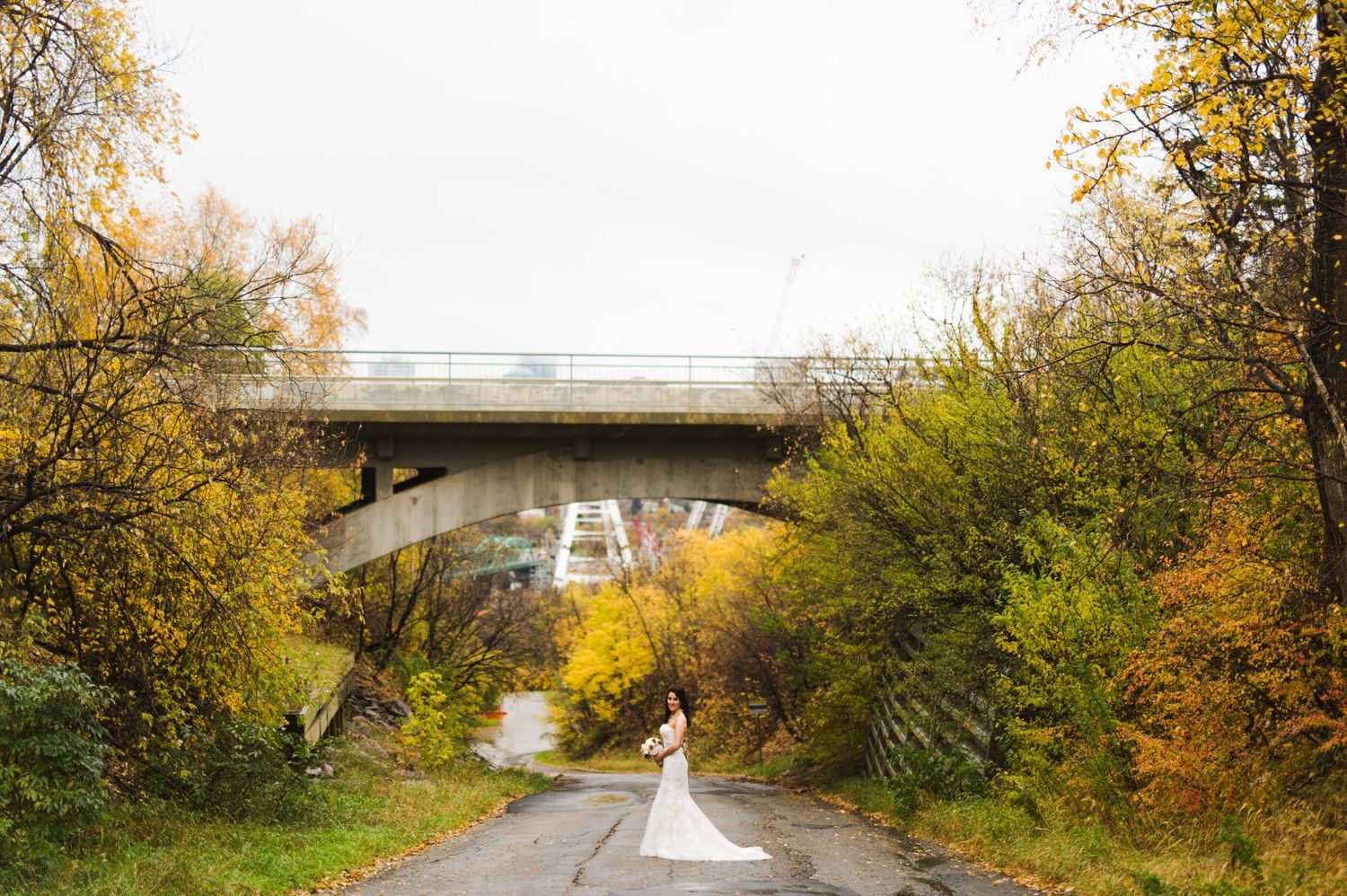 Bride walking down a tree-lined path toward an overpass, train tracks and cars above, surrounded by rich fall colors.