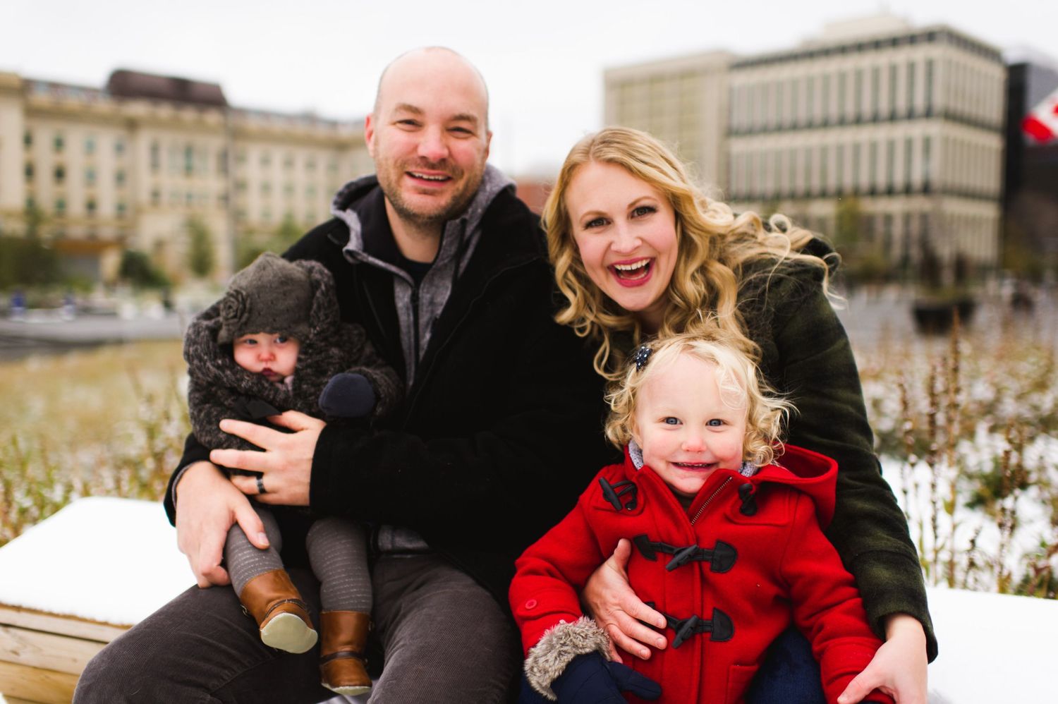 Smiling family of four sitting on a bench in a city park during winter, with two young children bundled in coats. Lifestyle family photo