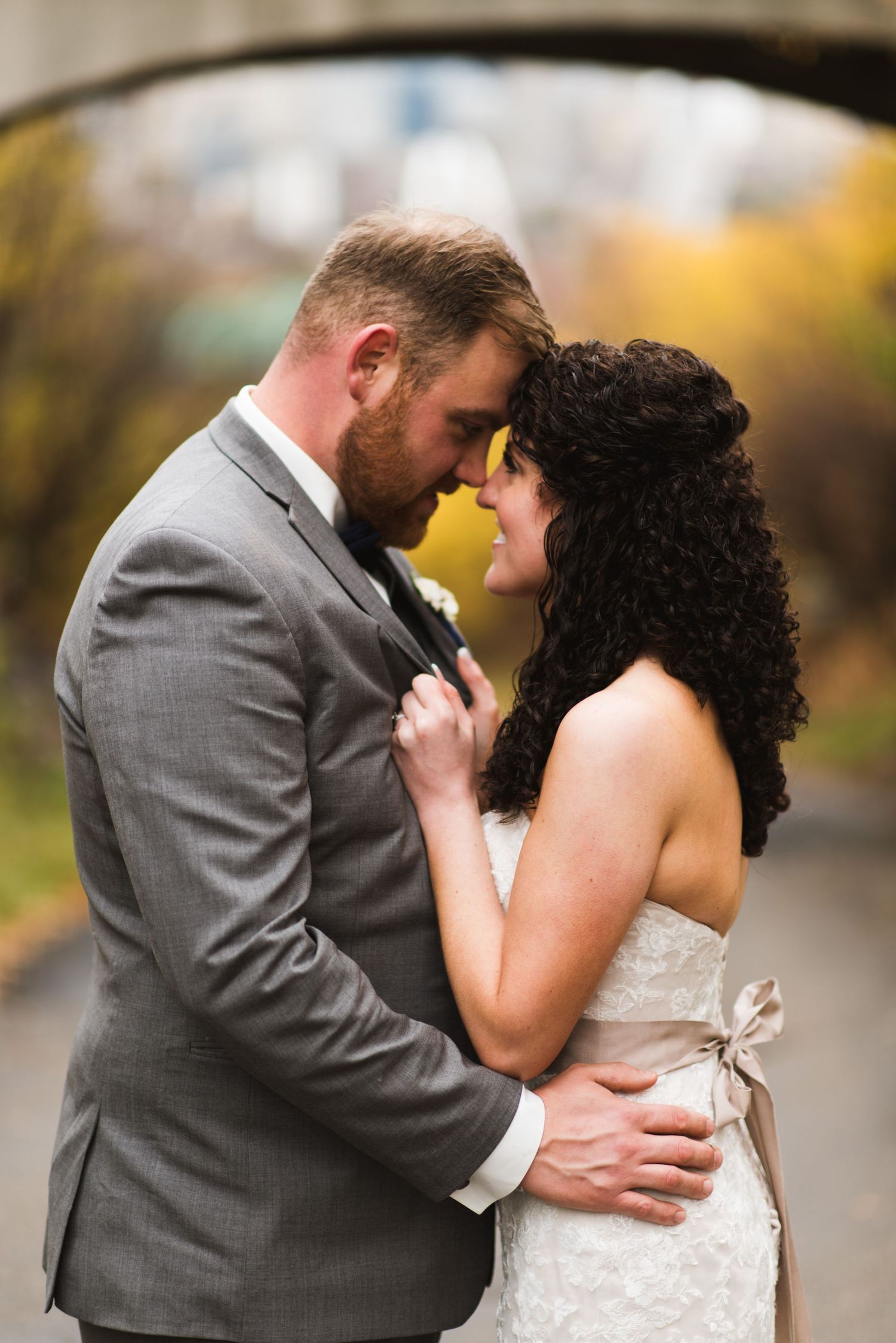 Bride and groom sharing an emotional forehead touch on a quiet city pathway with trees and an urban bridge behind them.