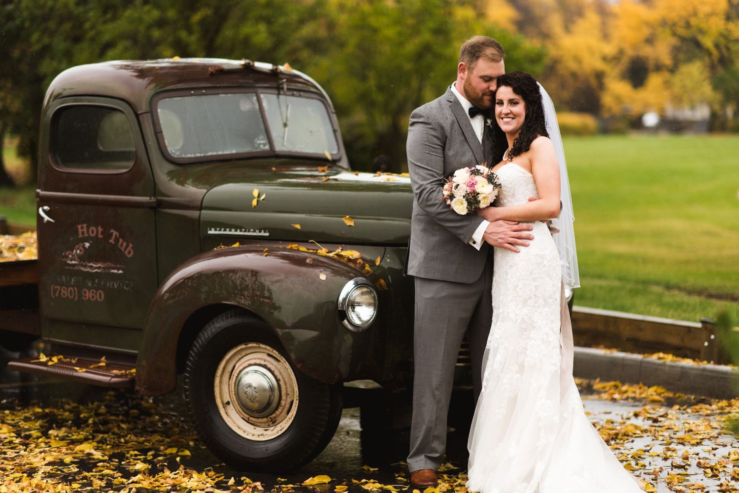 Bride and groom smiling beside a vintage green truck, standing on fallen autumn leaves.