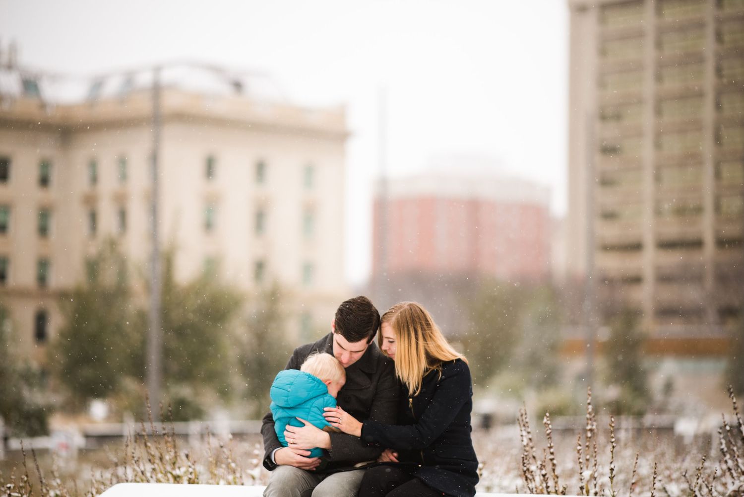 Urban family session downtown Calgary. Parents cuddle toddler on rooftop with city skyline in background.
