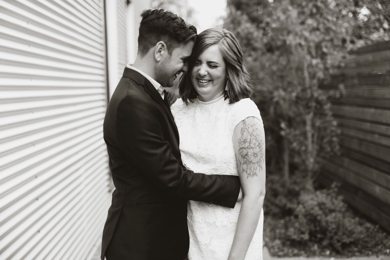 Candid black and white elopement photo; tattooed bride laughing with groom outside modern siding wall.