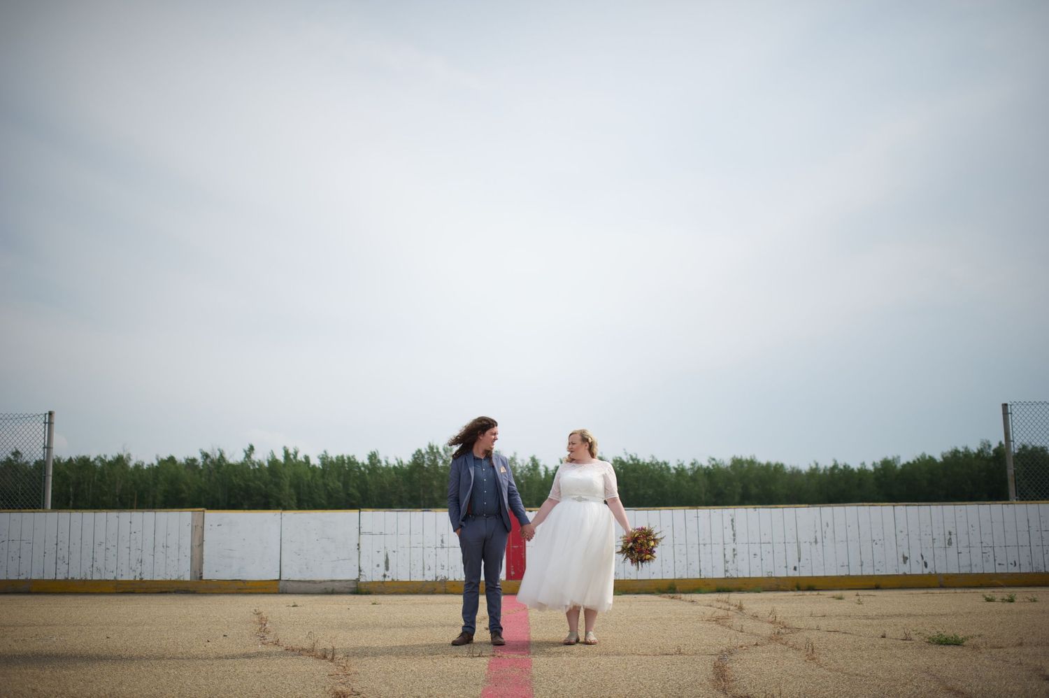 Newlyweds standing hand-in-hand on the center red line of an outdoor hockey rink, surrounded by tall trees and open sky.