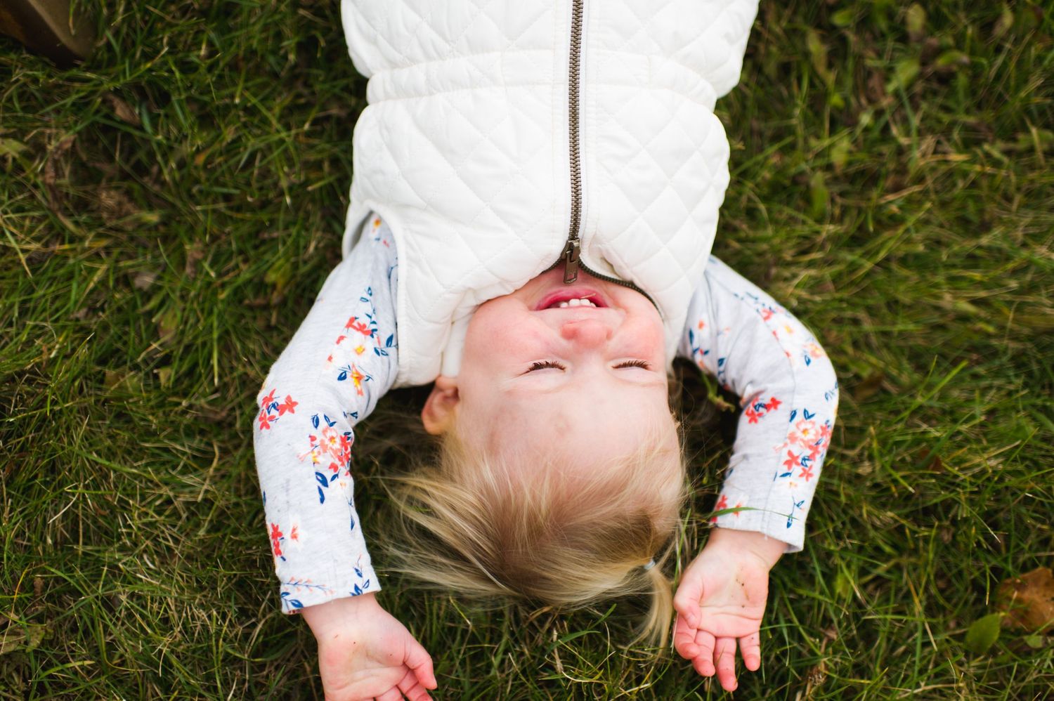 Blonde toddler in a white vest lying upside down in the grass, laughing with eyes closed and hands thrown back — playful and natural.