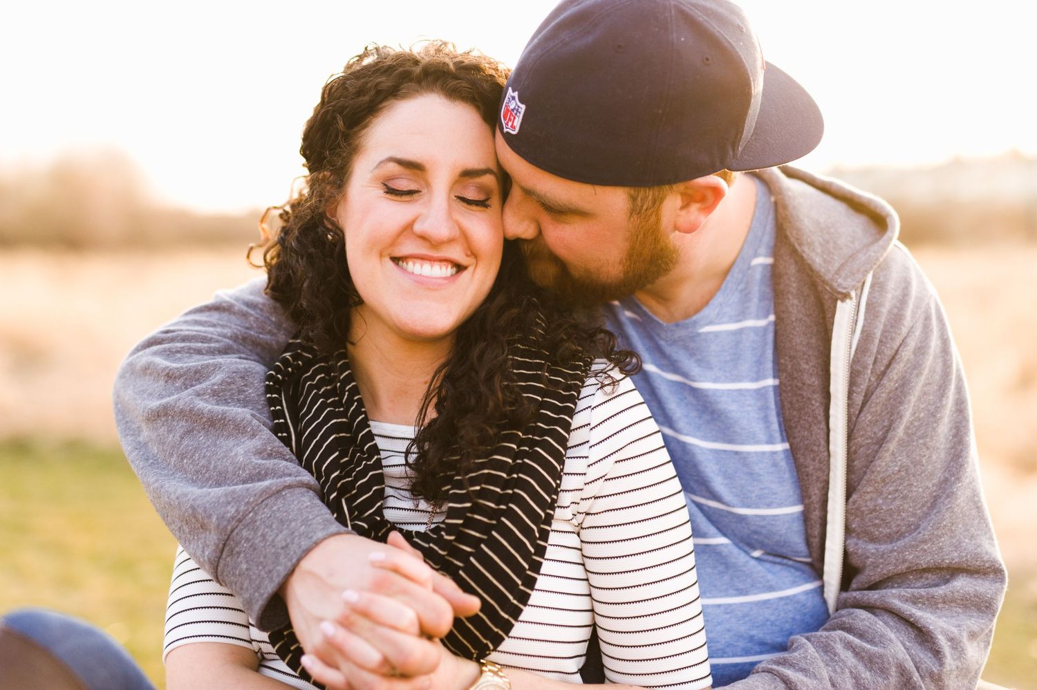 Warm sunset photo of a couple cuddling in a field, woman smiling while man kisses her cheek, dressed casually in fall layers.