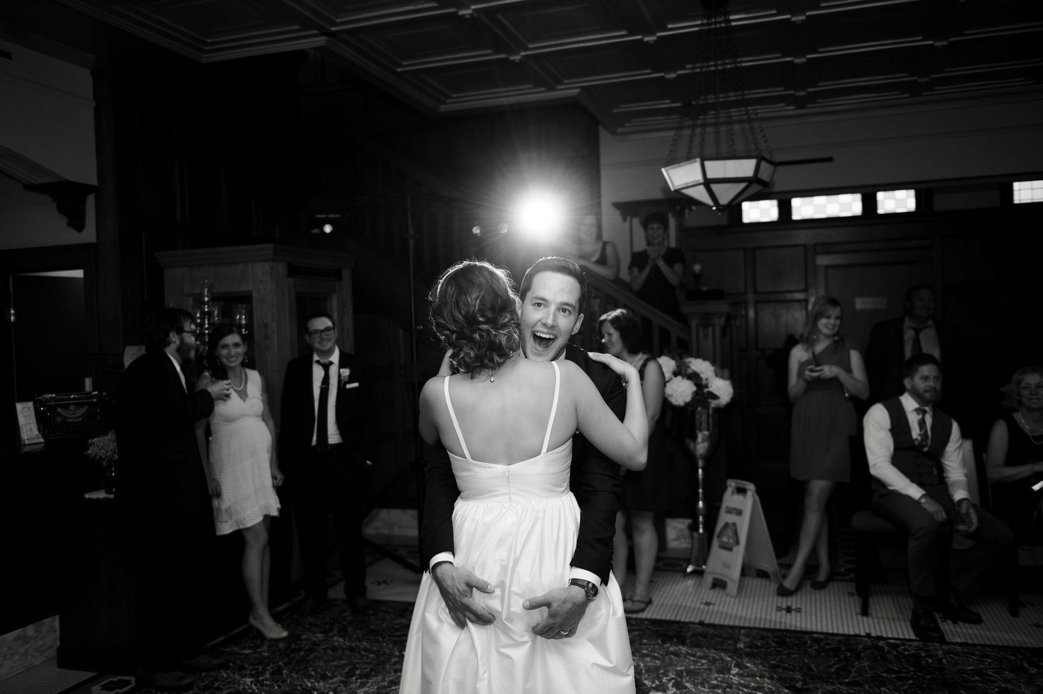 Black-and-white photo of a bride and groom dancing their first dance in a room full of watching guests.