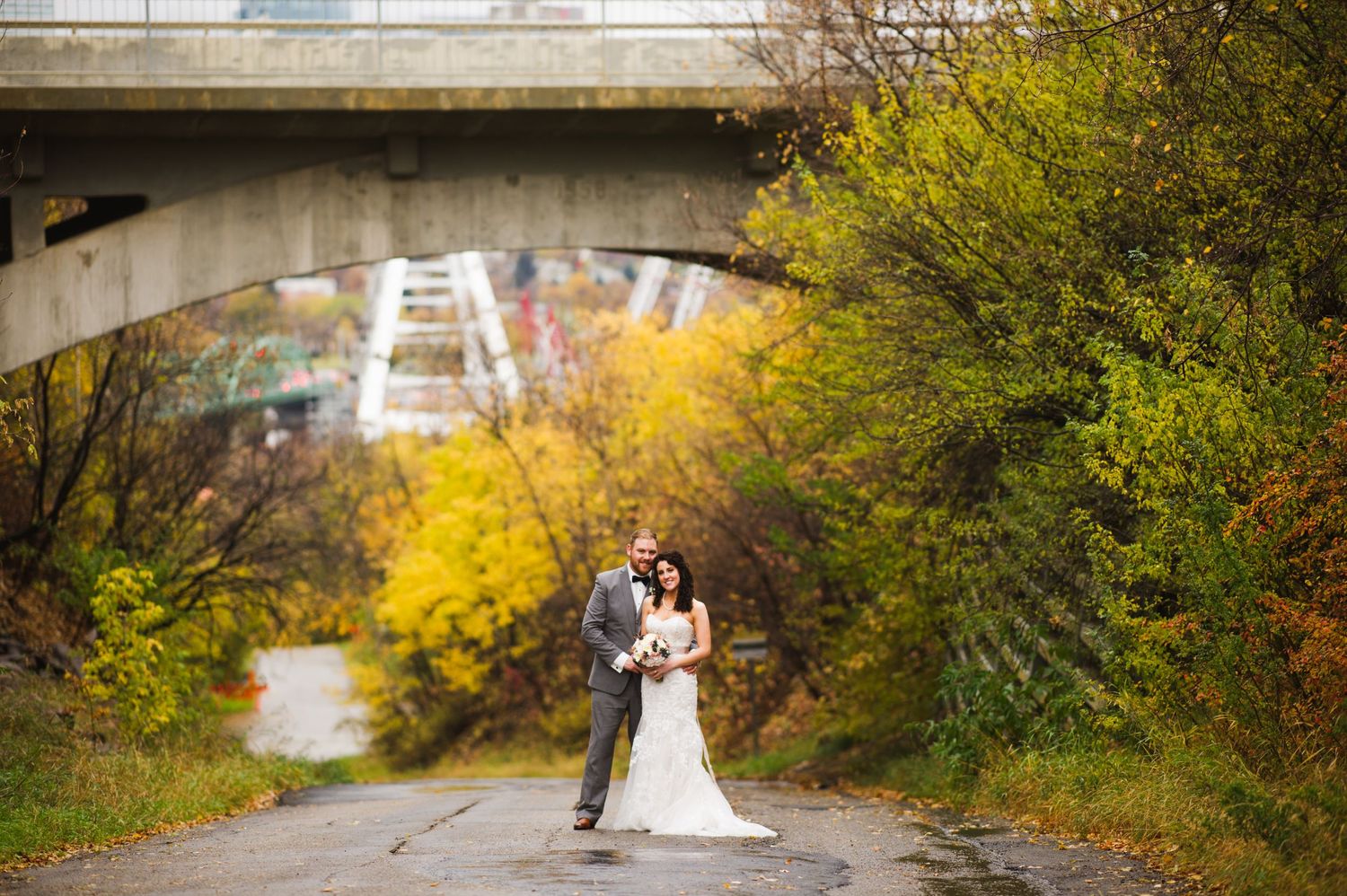 Bride and groom embracing on a quiet path beneath a bridge, surrounded by golden autumn trees and a distant view of the city skyline.