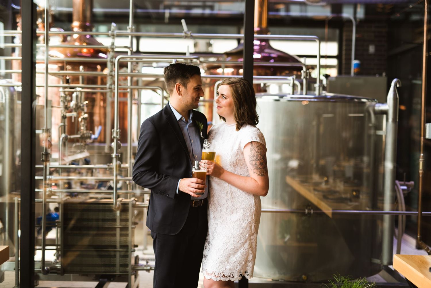 Industrial brewery wedding photo with couple holding drinks in front of tanks. Intimate Alberta micro-wedding.