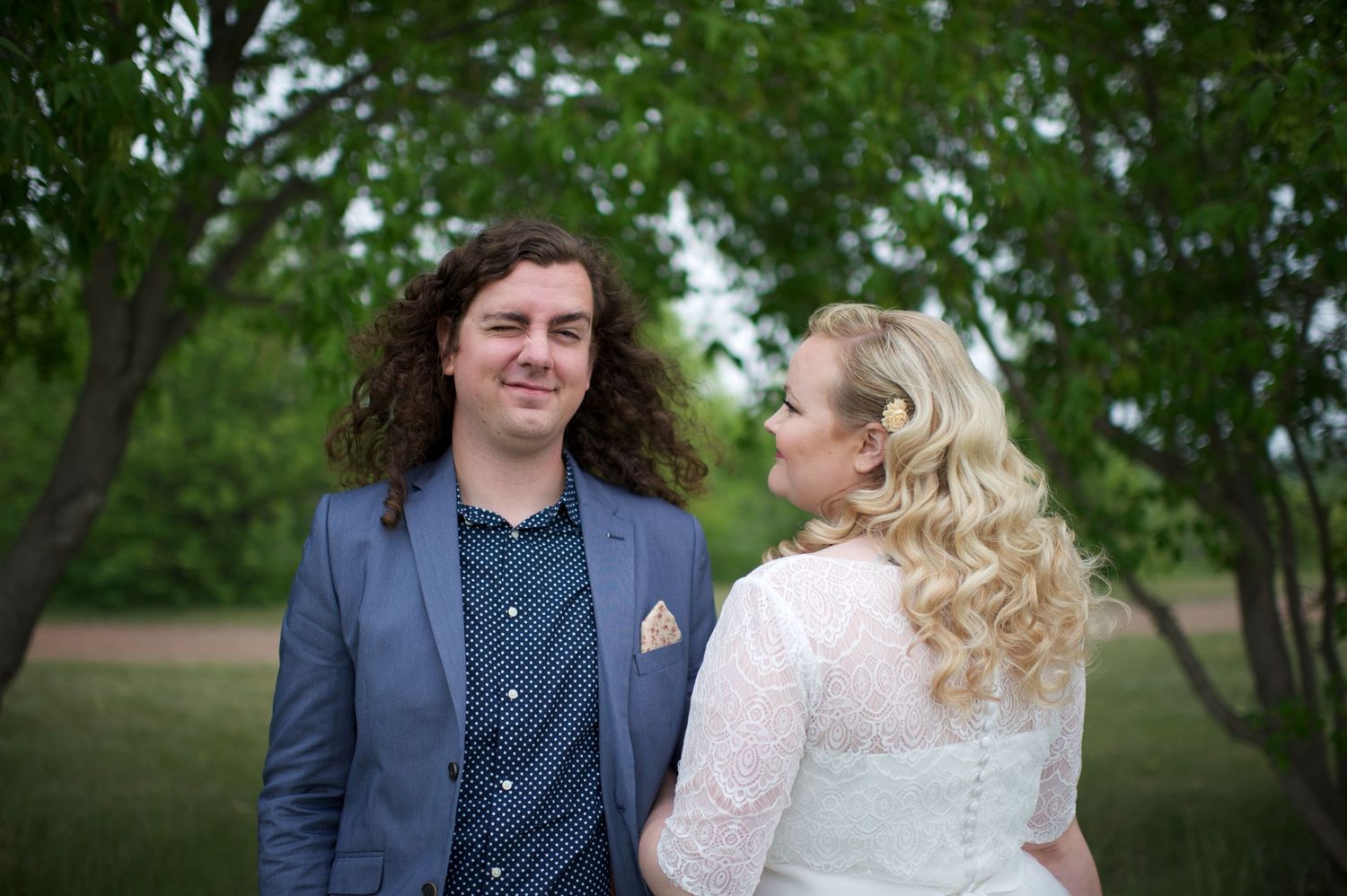 Wedding couple standing close together in a green park, bride looking lovingly at groom while he faces the camera with a soft smile.
