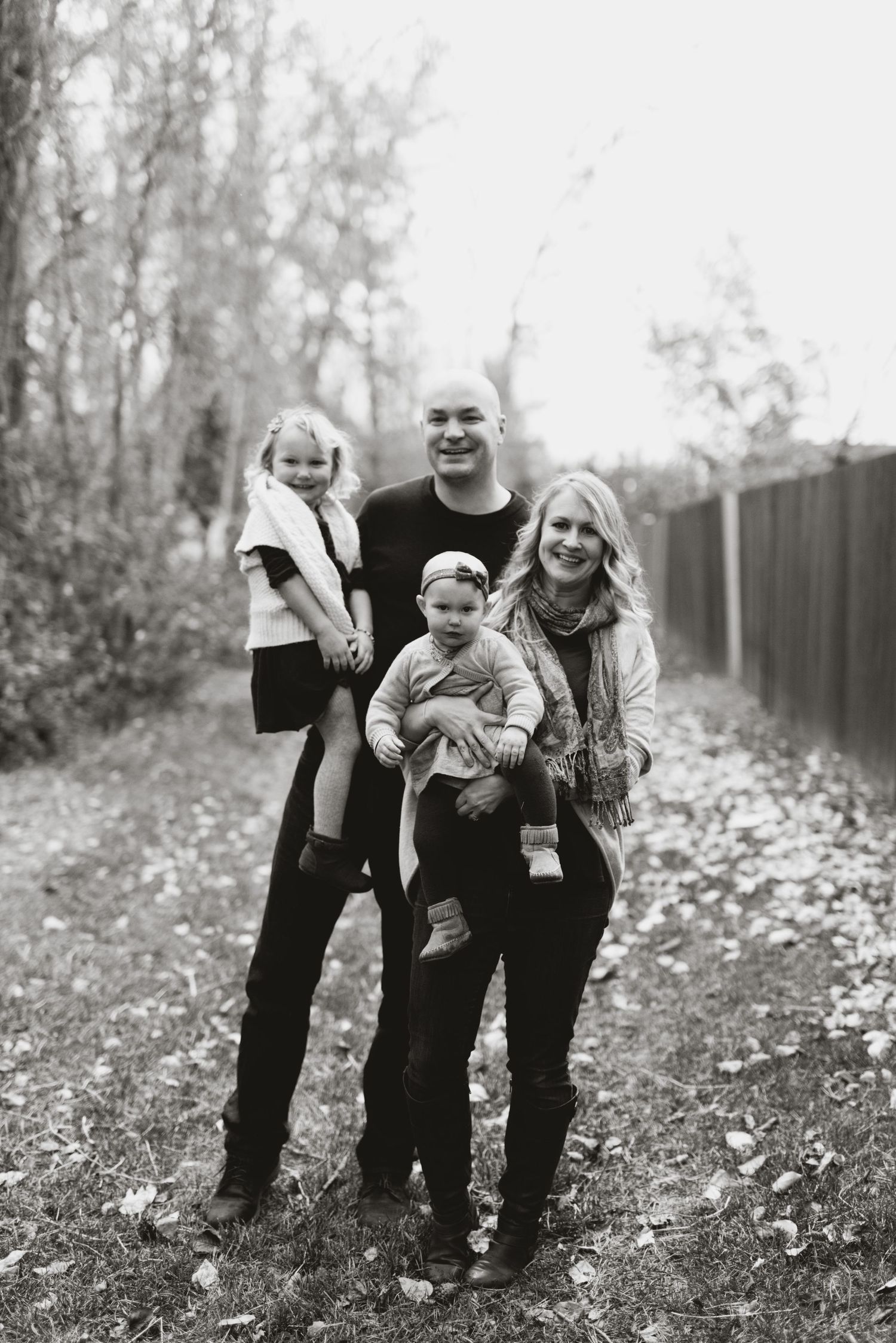Black and white vertical portrait of a young family with two kids on a leaf-covered pathway, surrounded by trees and a wooden fence.