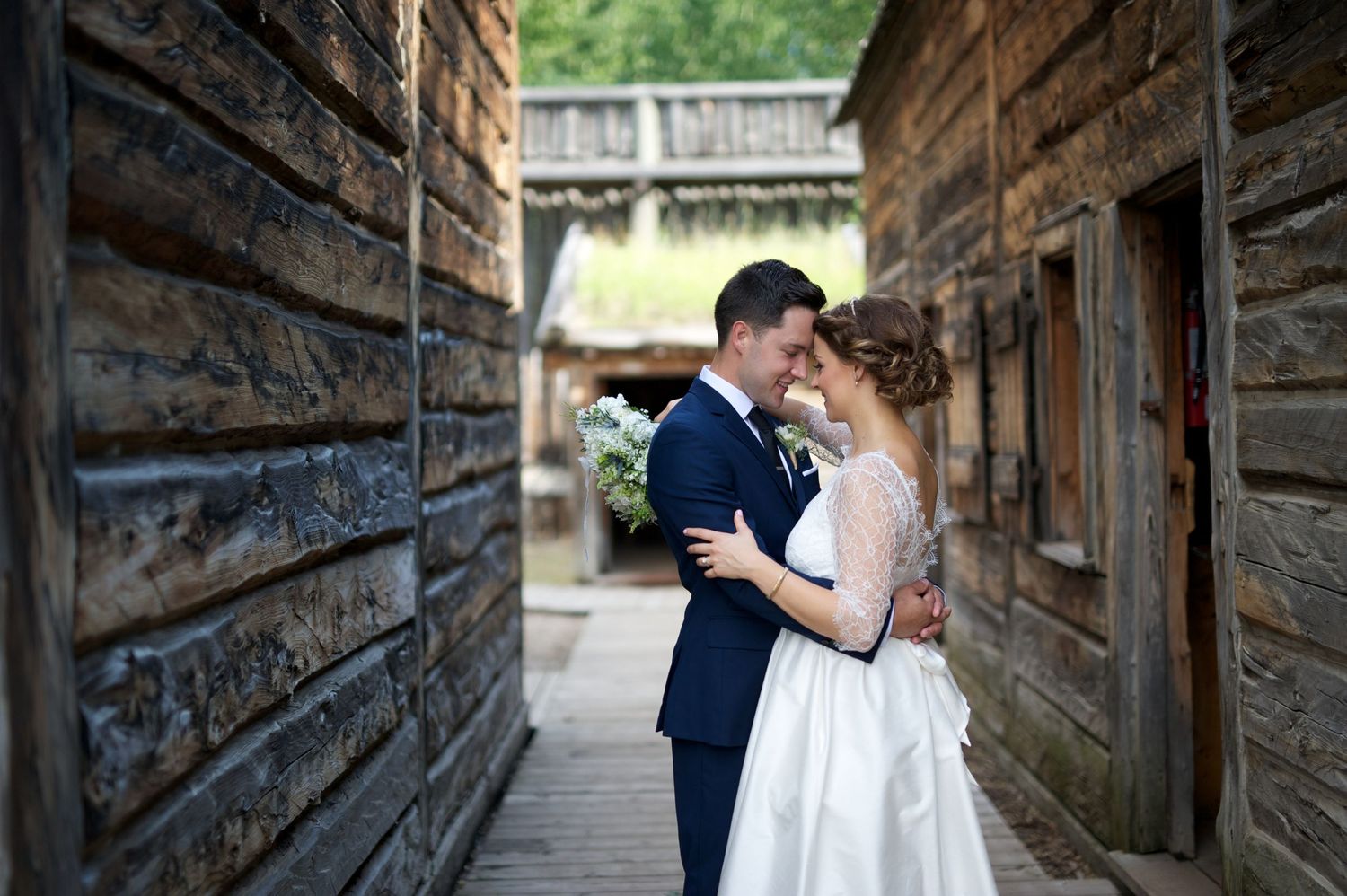 Bride and groom forehead-to-forehead between rustic wood buildings, sharing an intimate moment.