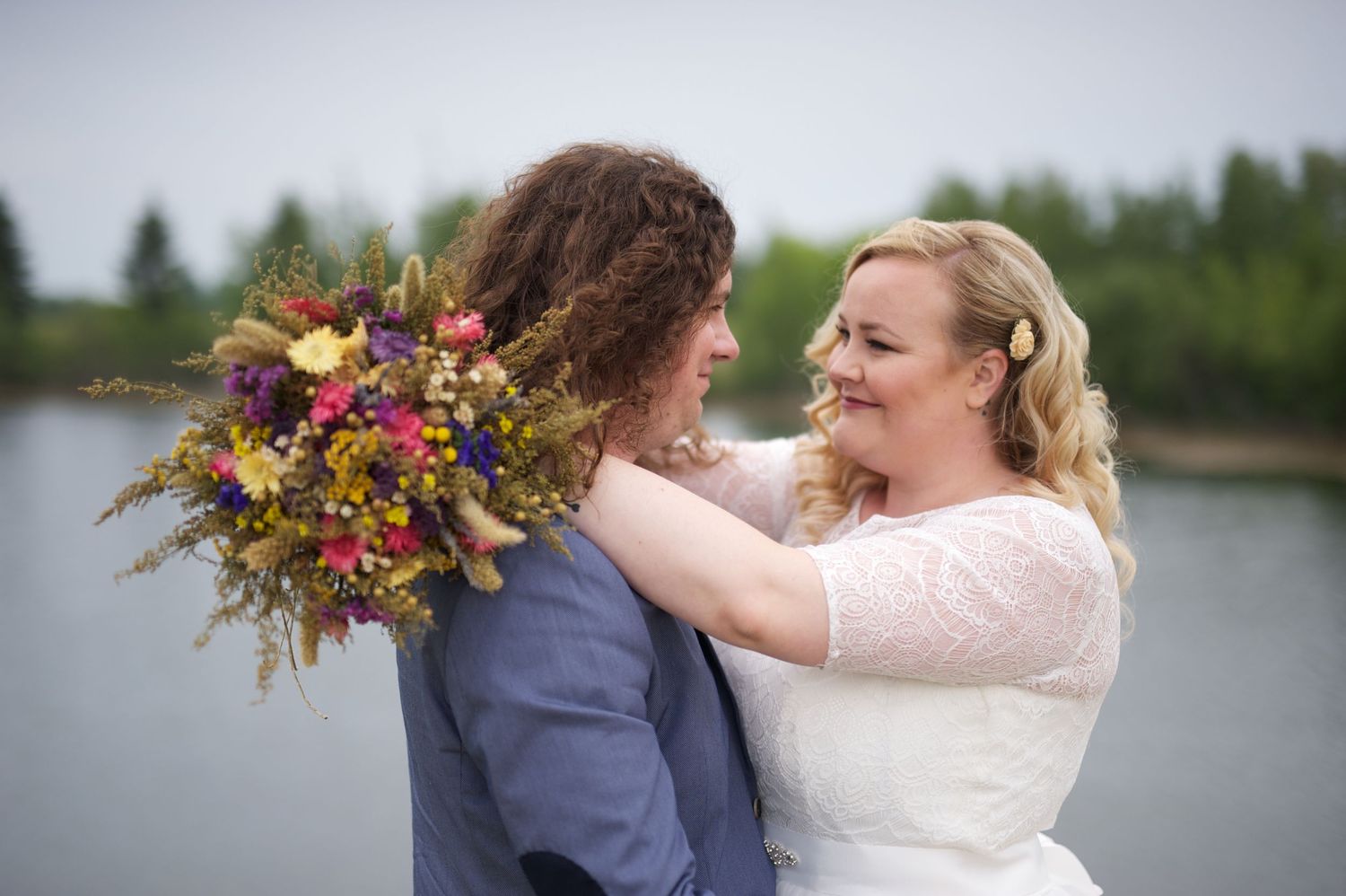 Bride embracing her groom by a quiet pond, bouquet of wildflowers over his shoulder, both smiling with soft joy.