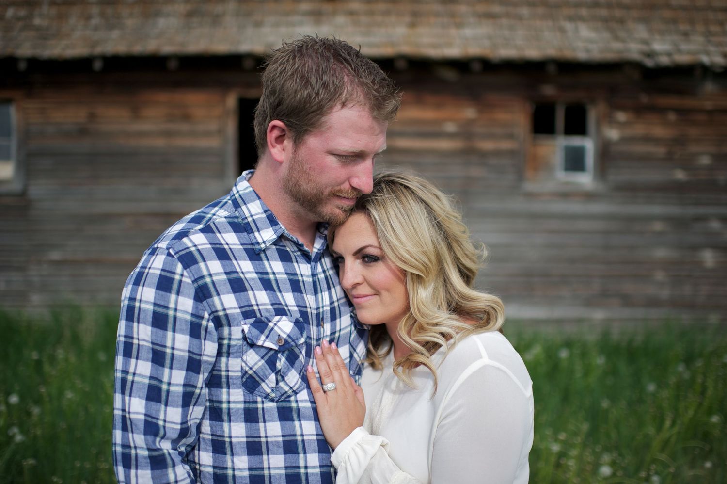 Engaged couple standing close in a grassy field with a rustic barn behind them, the woman resting her head on her partner’s chest.