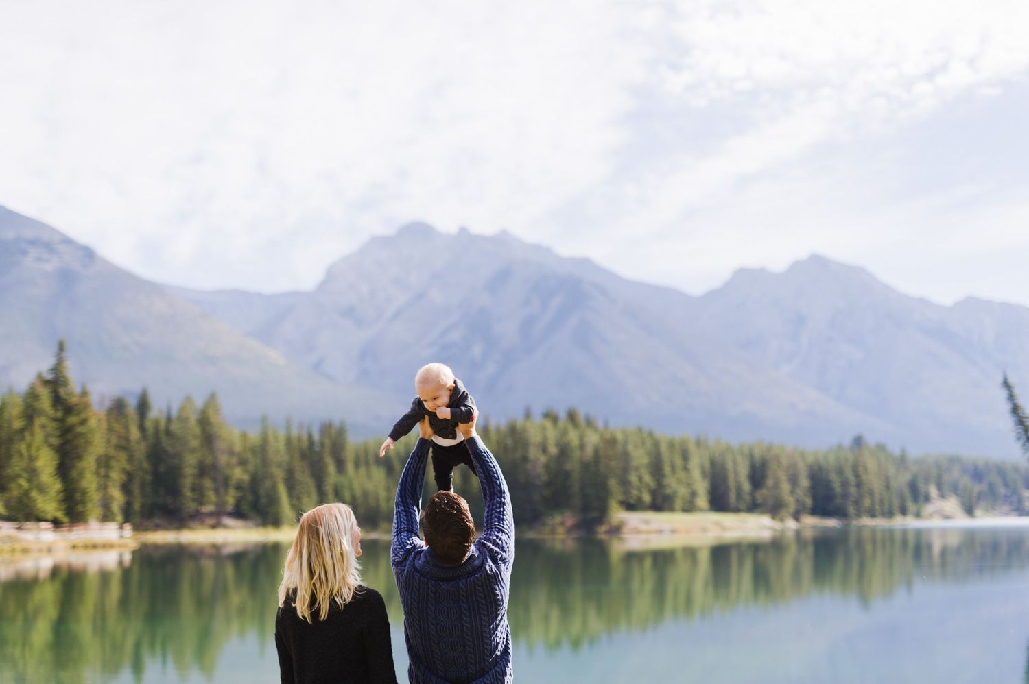 Dad lifts baby high above lake with mountains behind. Alberta adventure family session in the Rockies.