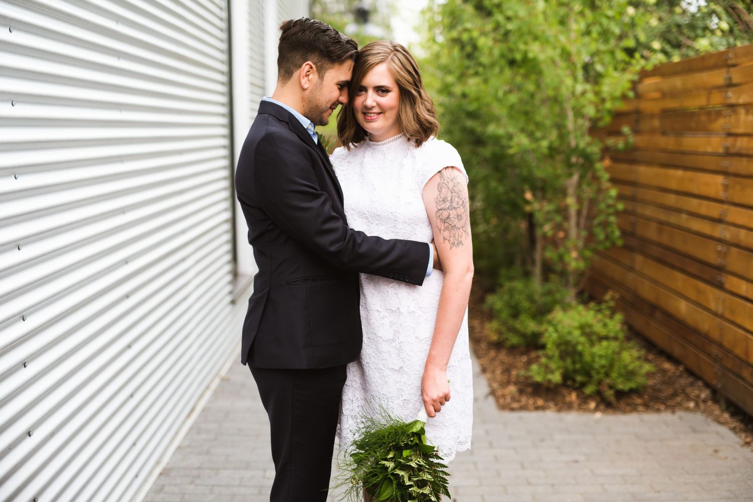 Relaxed wedding portrait beside corrugated siding and wooden fence. Tattooed bride holding bouquet during Calgary elopement.