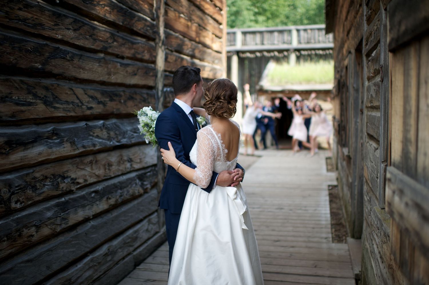 Bride and groom share a quiet moment, arms around each other, as wedding guests gather in the background of a heritage site.