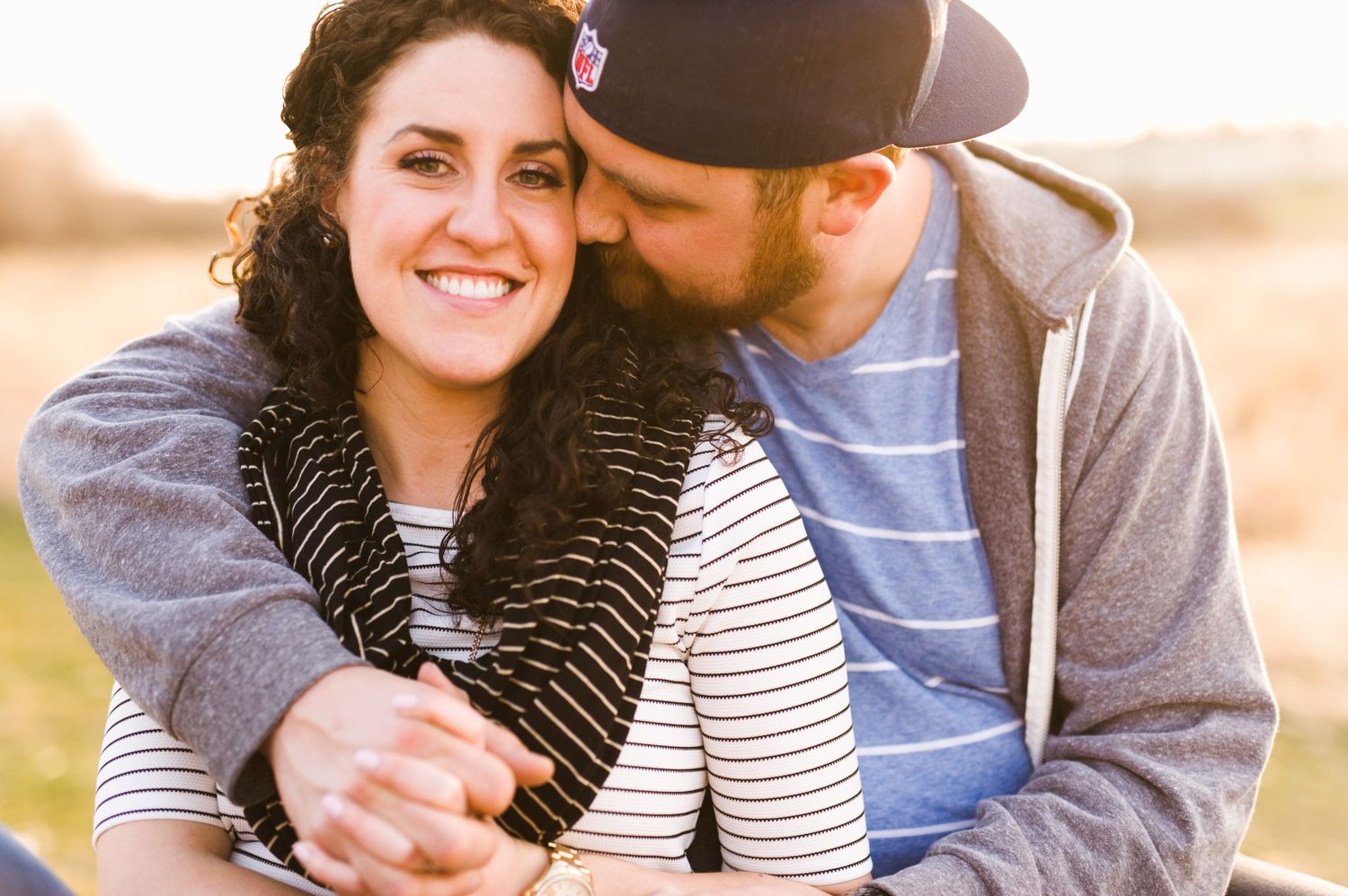 Warm sunset photo of a couple cuddling in a field, woman smiling while man kisses her cheek, dressed casually in fall layers.