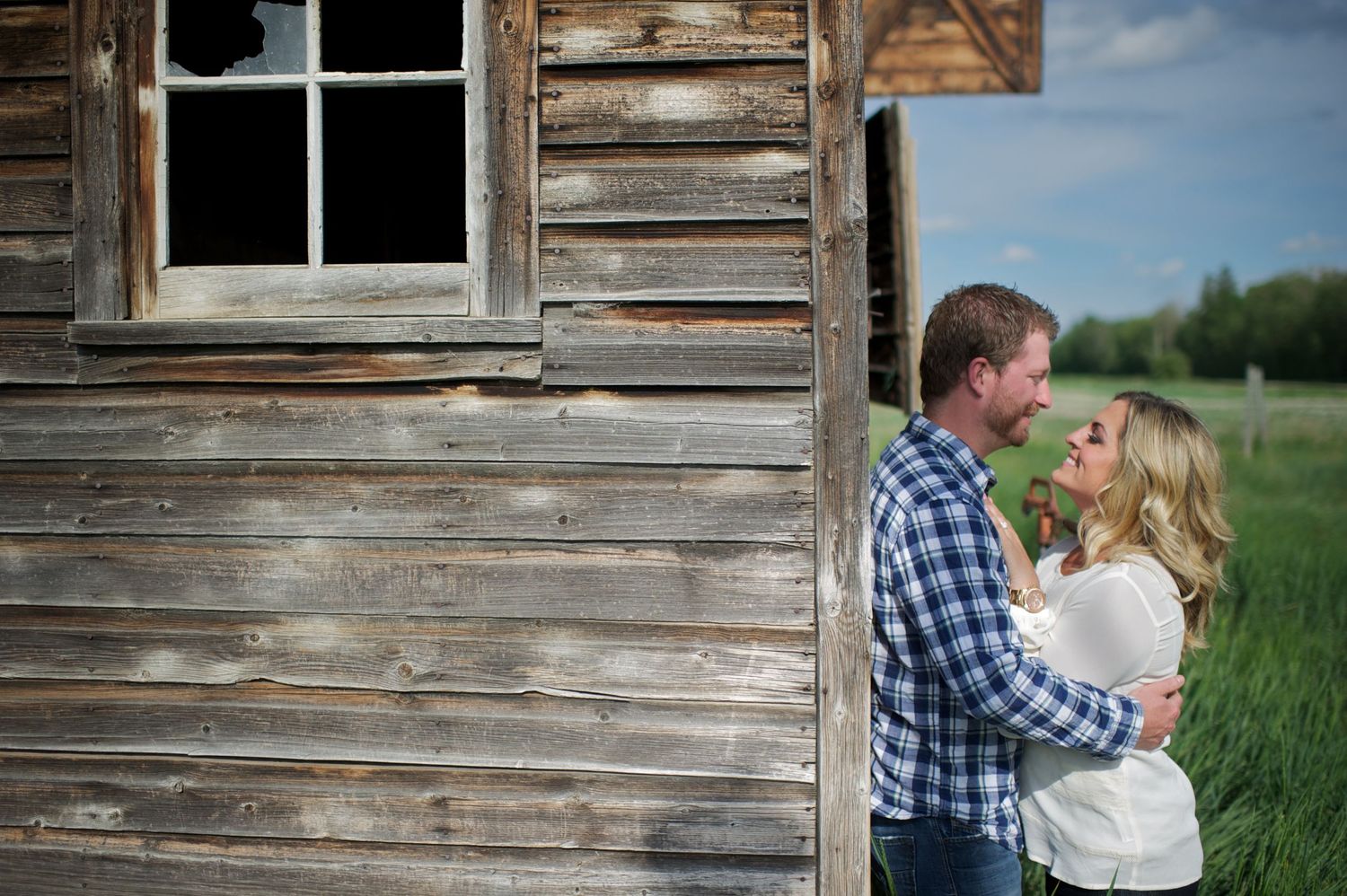 Romantic photo of a couple standing face to face beside a weathered wooden cabin, surrounded by green field and blue skies.