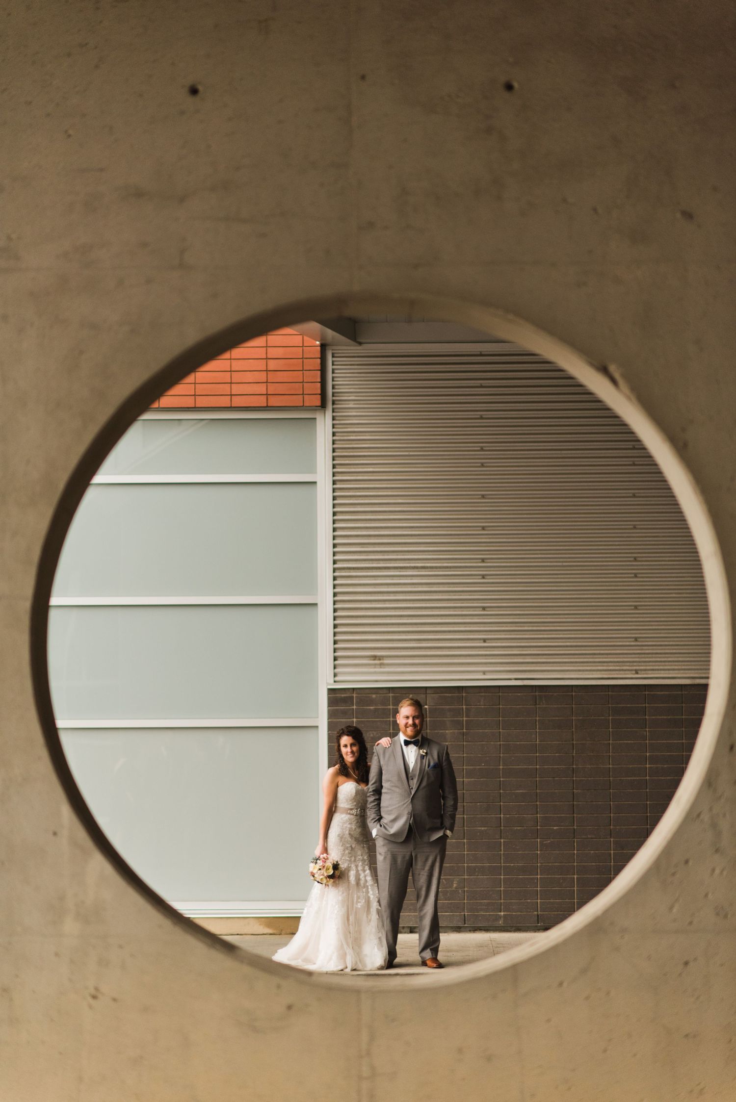 Wedding couple standing in an urban setting, perfectly framed by a modern concrete circular cutout — bold, artistic portrait with city vibe