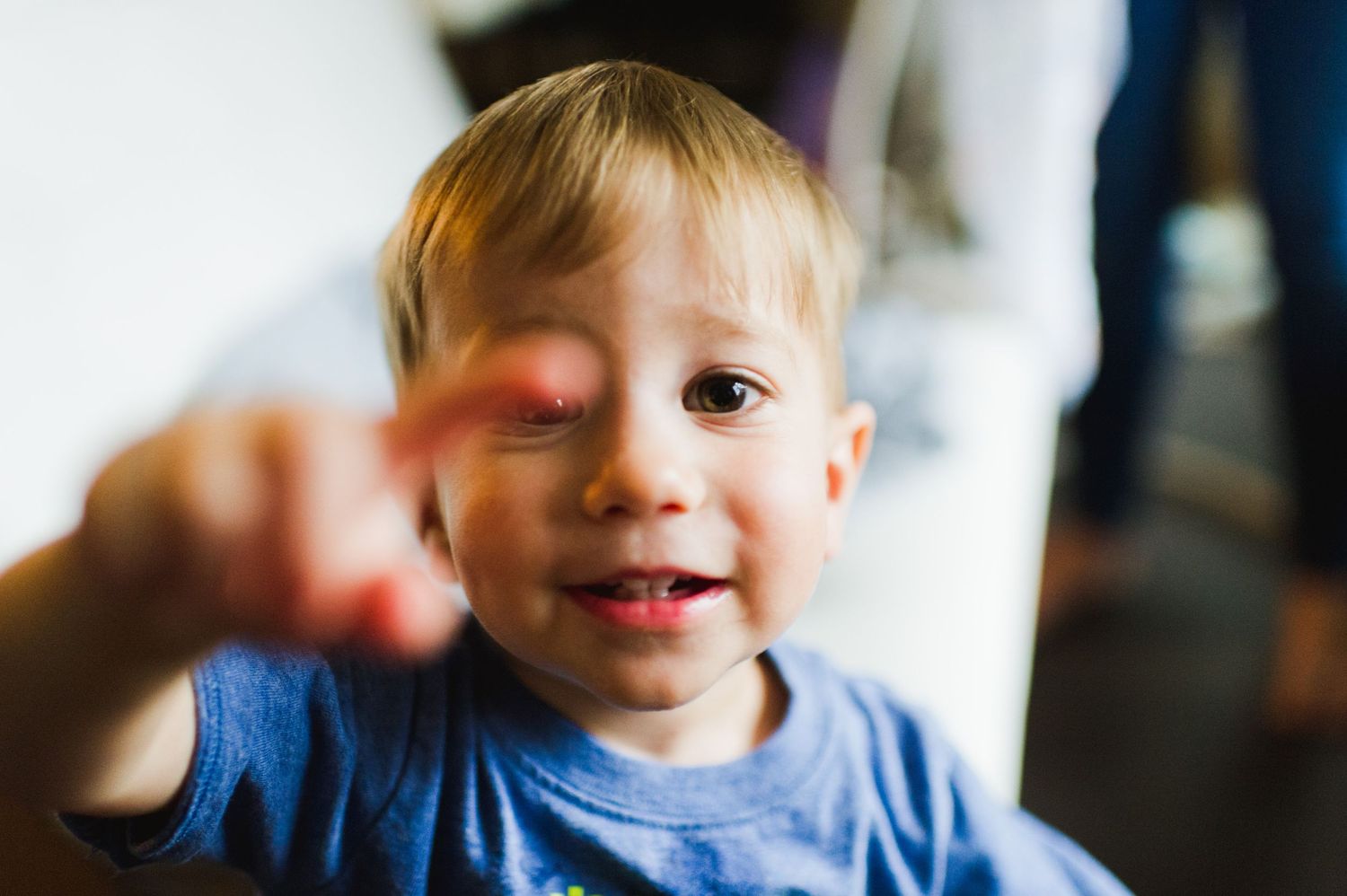 Toddler boy smiling and reaching toward the camera indoors, surrounded by blurred figures in the background. Playful lifestyle portrait.