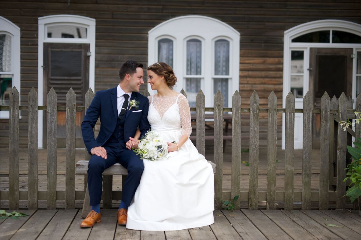 Bride and groom sit on a bench, smiling at each other, in front of a heritage building with white-trimmed windows.