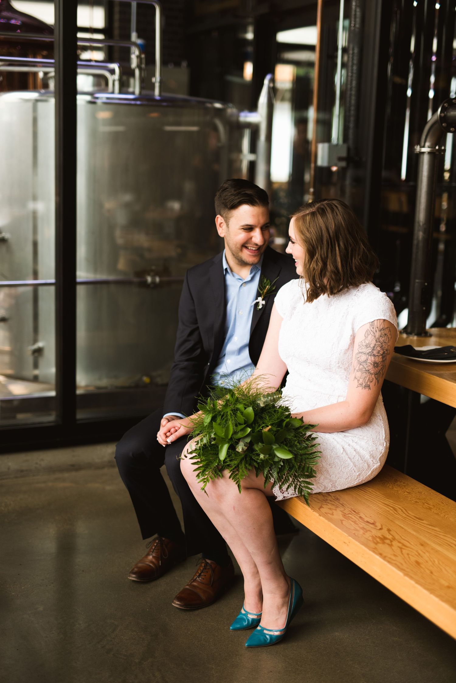 Brewery elopement couple sitting indoors. Bride in short dress and blue shoes holding bouquet. Industrial wedding vibes.
