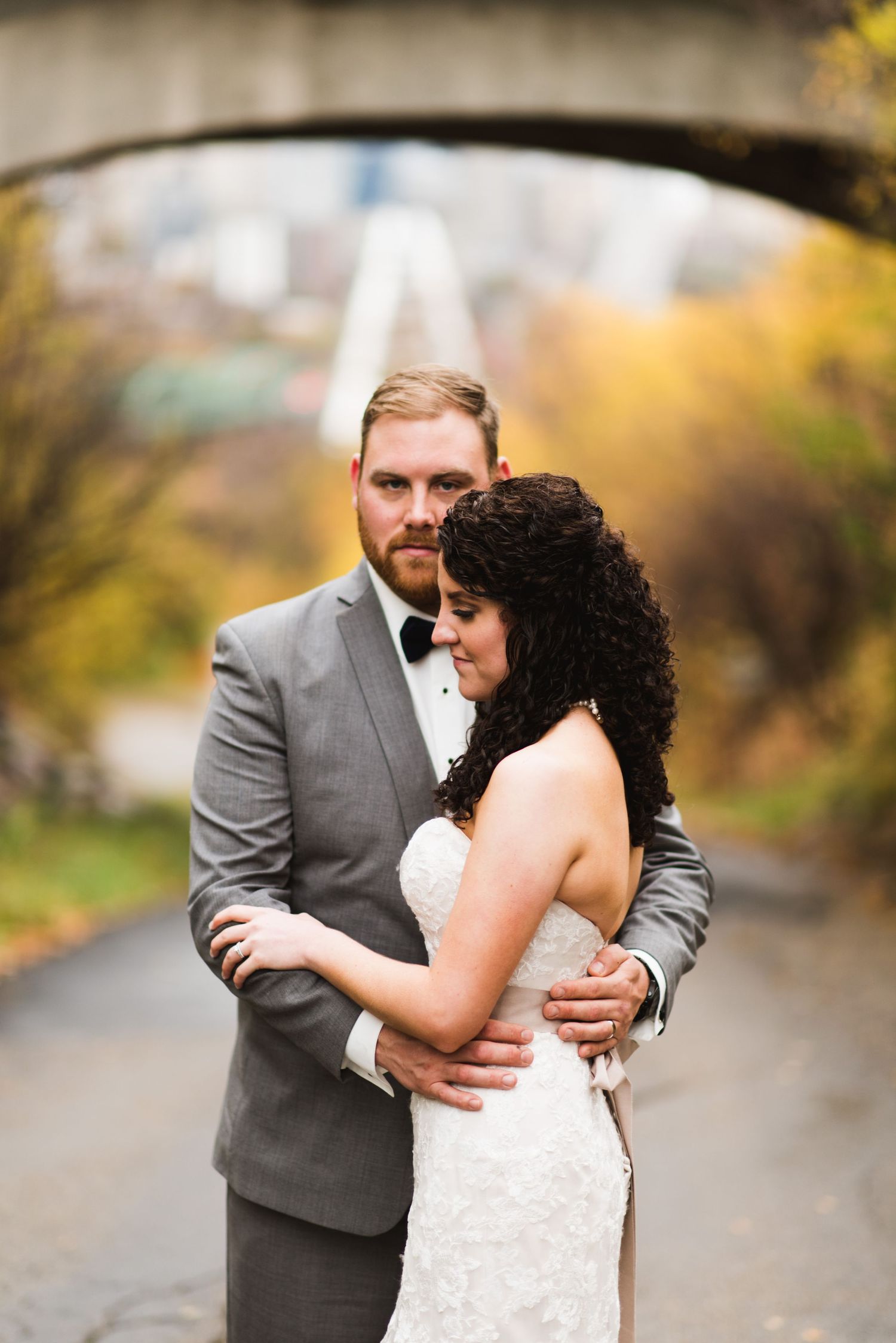 Bride and groom standing together under an overpass with the Calgary skyline blurred in the background, golden leaves framing the moment.