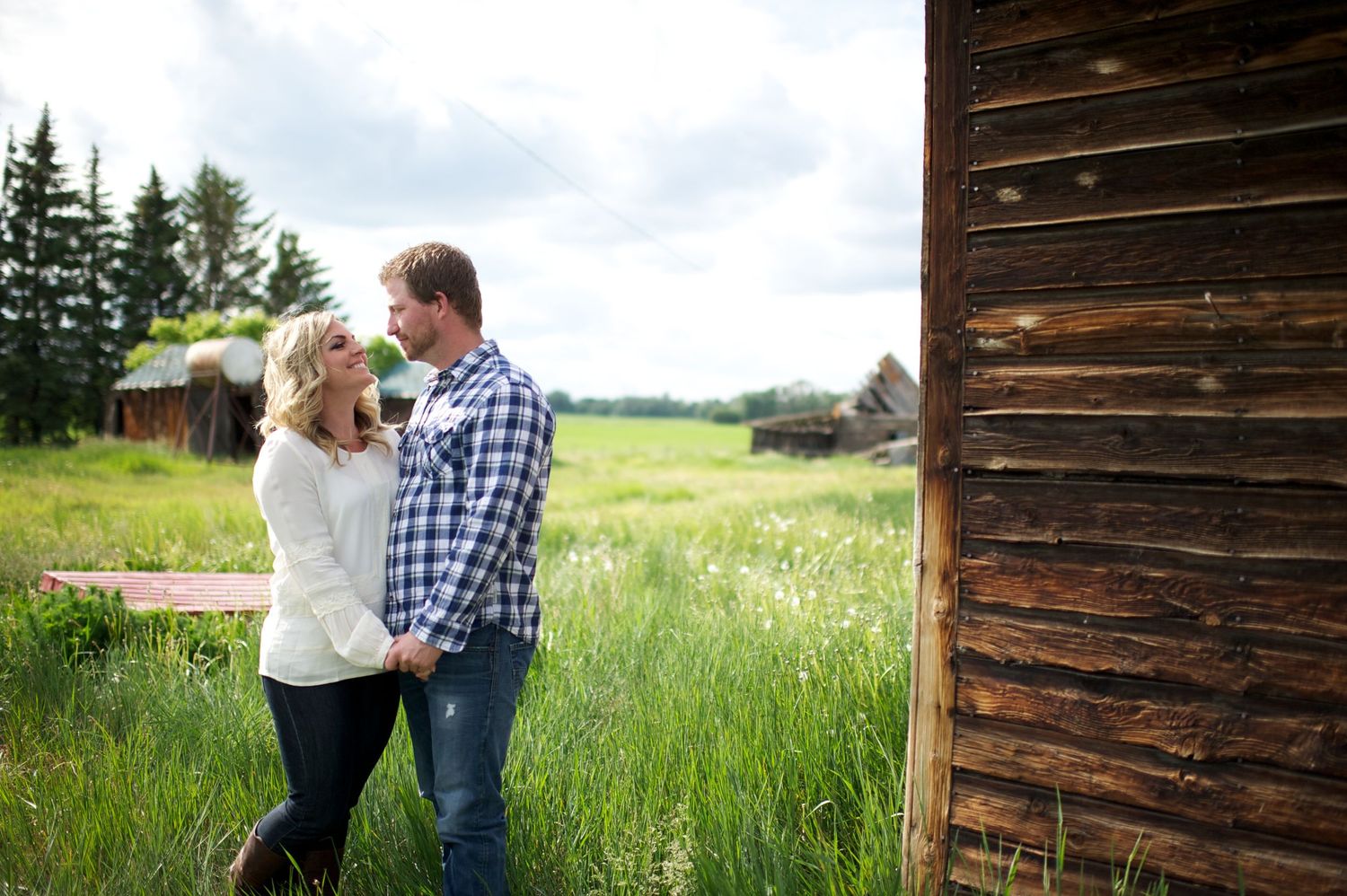 Romantic lifestyle photo of a couple standing in tall grass beside a weathered barn, holding hands and smiling at each other.