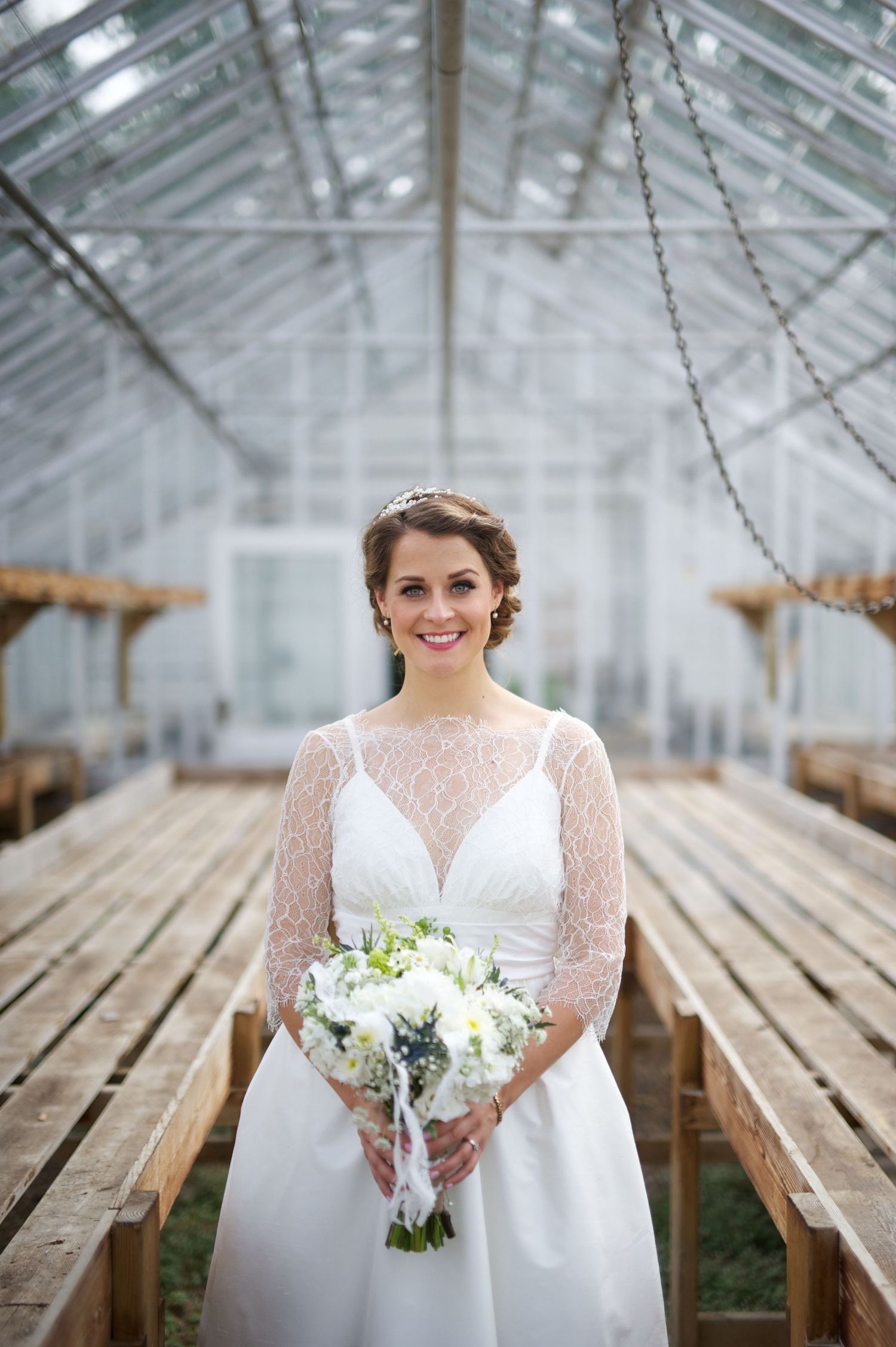 Bride stands centered in a greenhouse, holding her bouquet with a quiet smile. Natural, simple, and full of light.