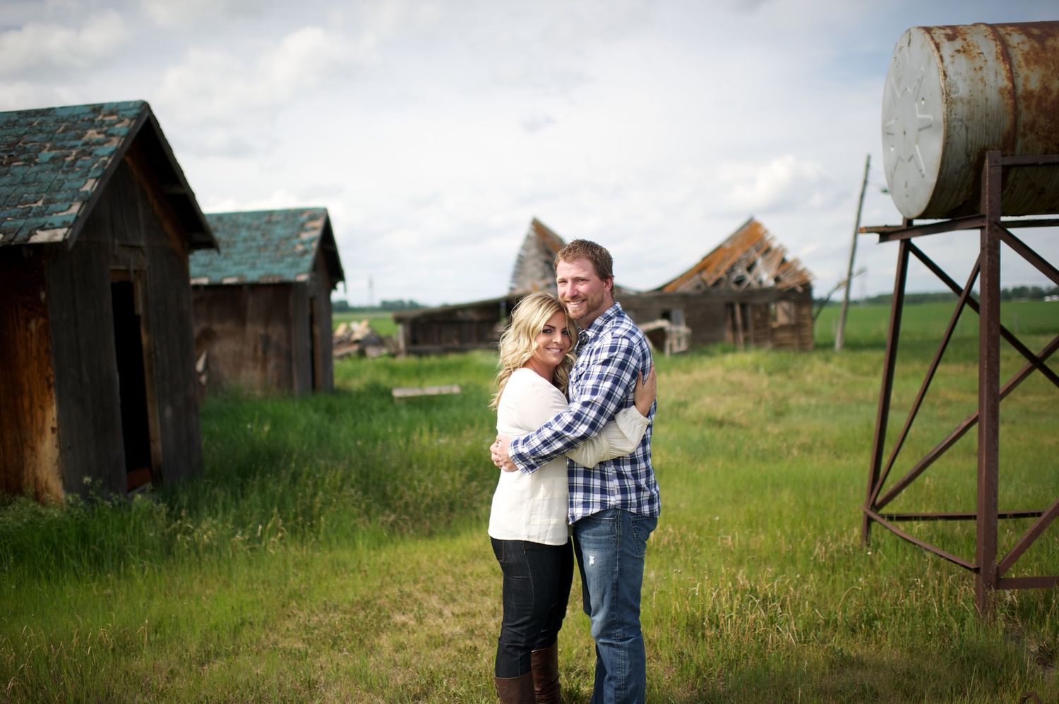 Engaged couple hugging in front of weathered farm buildings and an old water tank, surrounded by green grass and prairie landscape.