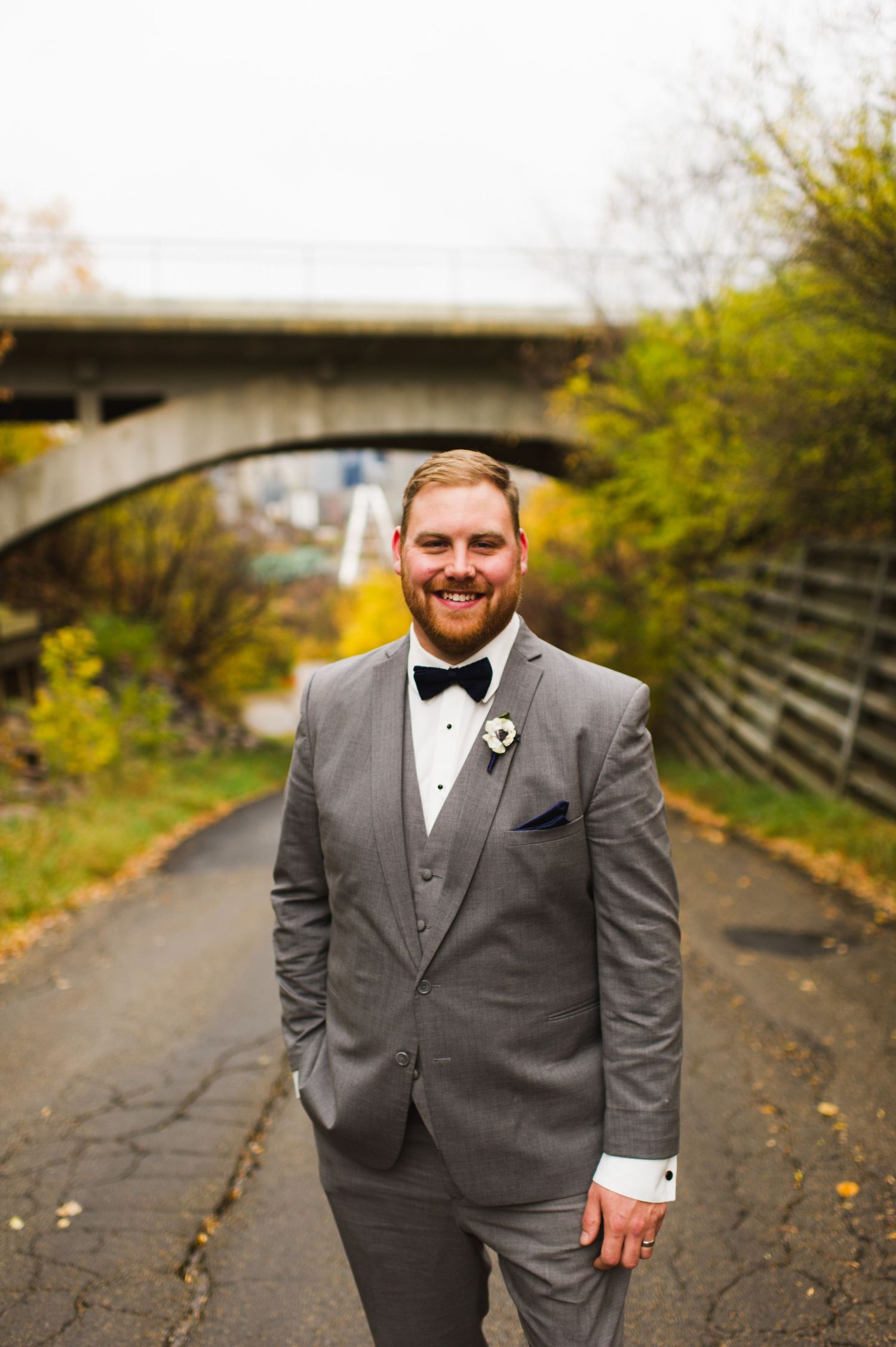 Groom standing confidently in a grey suit and bow tie on a tree-lined path with an urban overpass in the background, fall leaves scattered