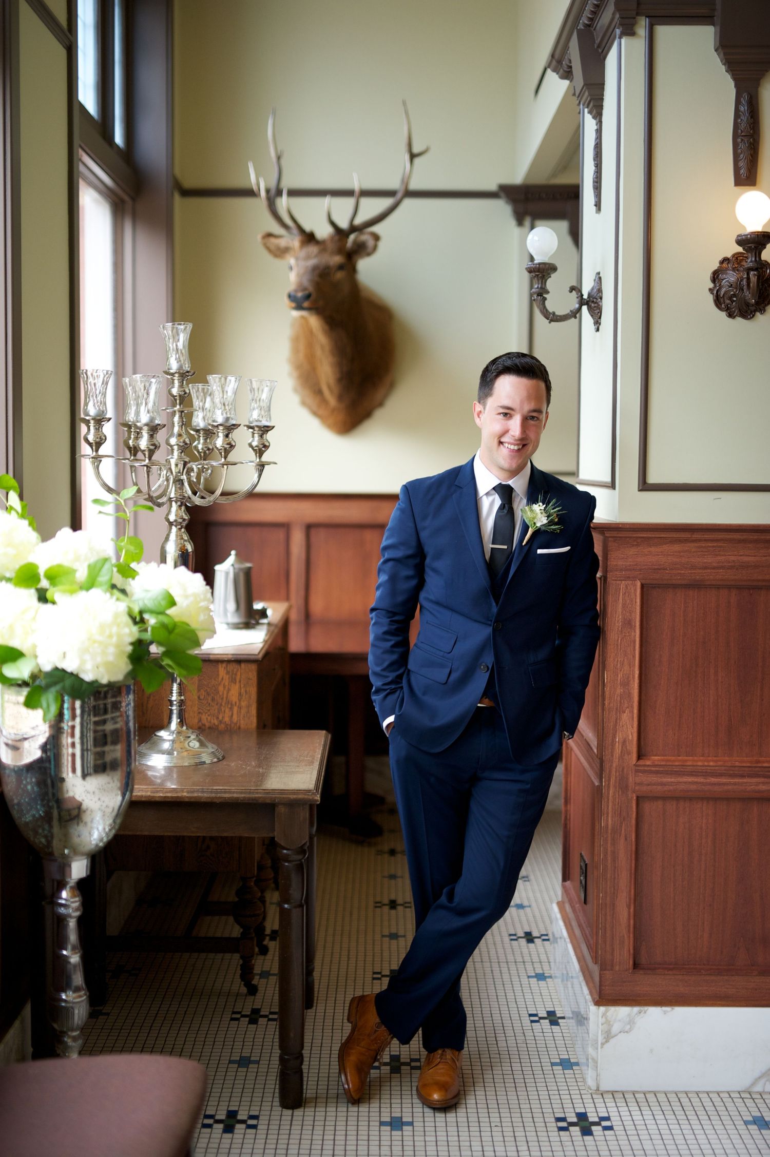 Groom leans casually against wood paneling, surrounded by vintage charm and soft floral accents.