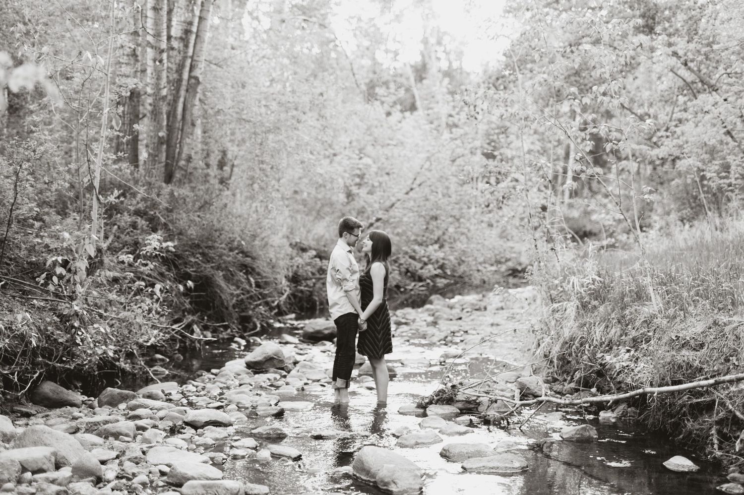 Black-and-white photo of a couple standing barefoot in a rocky creek, gently touching foreheads.