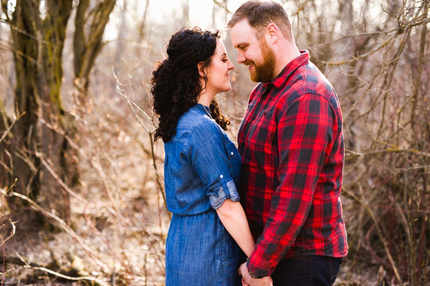 Couple holding hands and smiling softly at each other in a bare forest, with golden light highlighting their faces and tender expressions.