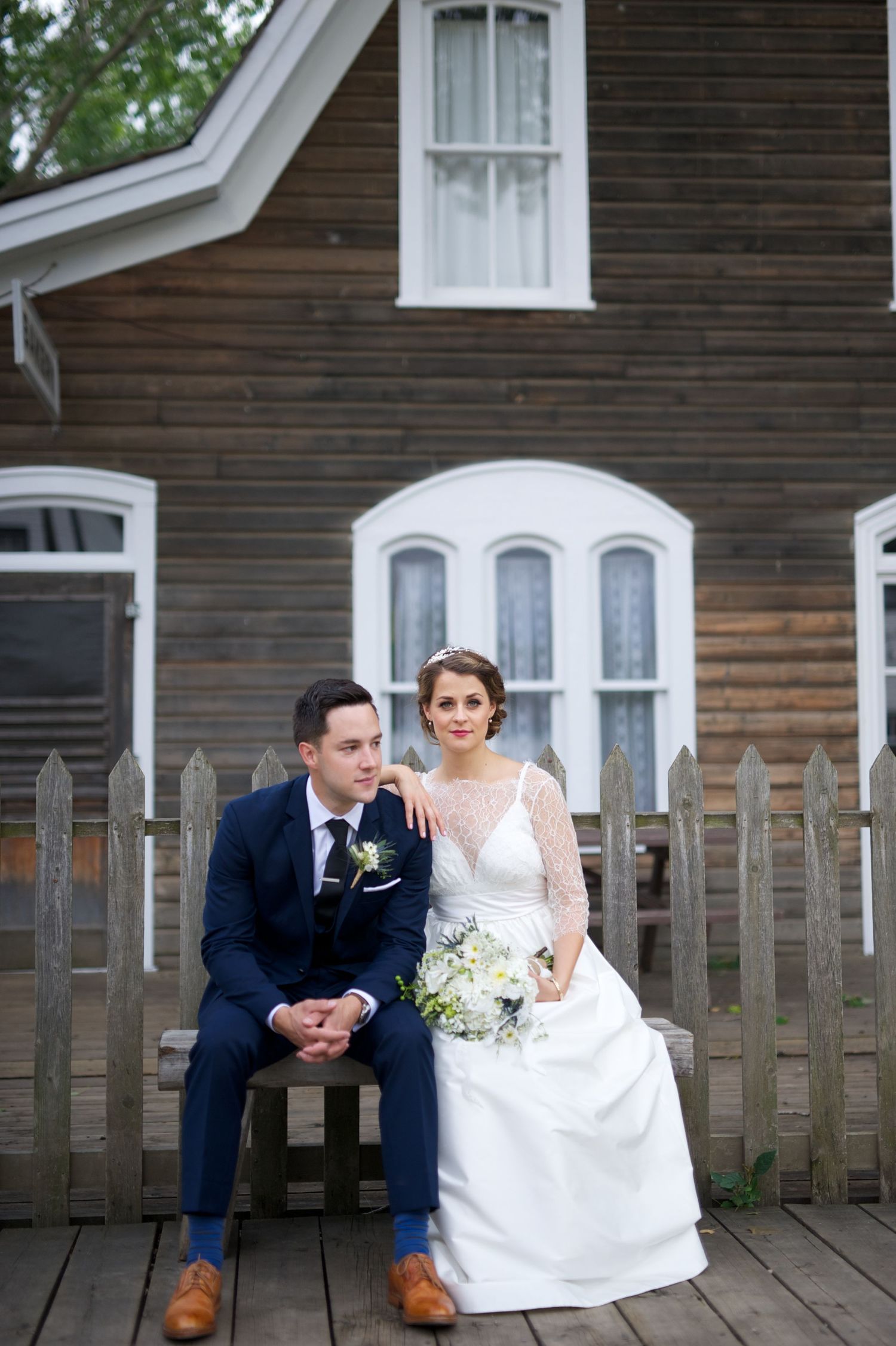 Newlyweds sit on a wooden bench in front of a heritage house, holding hands and smiling calmly.