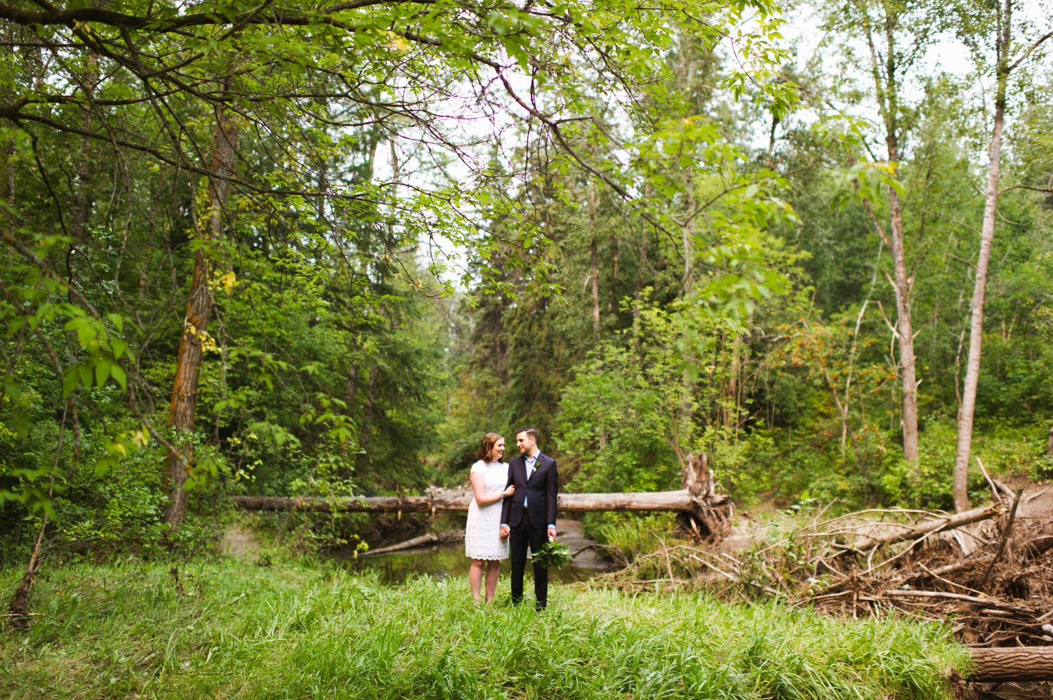 Full-body wide shot of couple surrounded by fallen trees and tall grass. Edmonton elopement with woodland vibes.