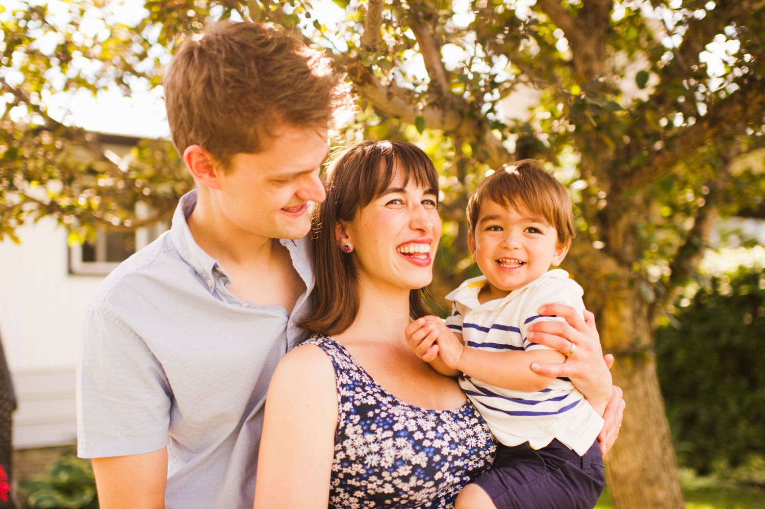 Young couple smiling while holding their toddler, all laughing under a leafy backyard tree.