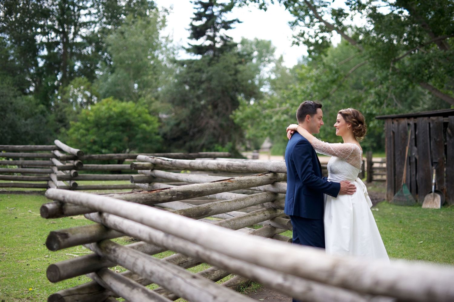Bride and groom embrace and smile beside a rustic split-rail fence, surrounded by lush trees.