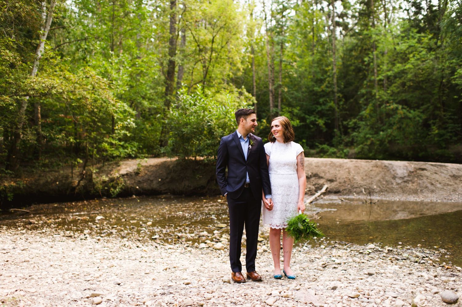 Bride and groom pause together by the riverbank. Nature elopement in Alberta with forest and water backdrop.