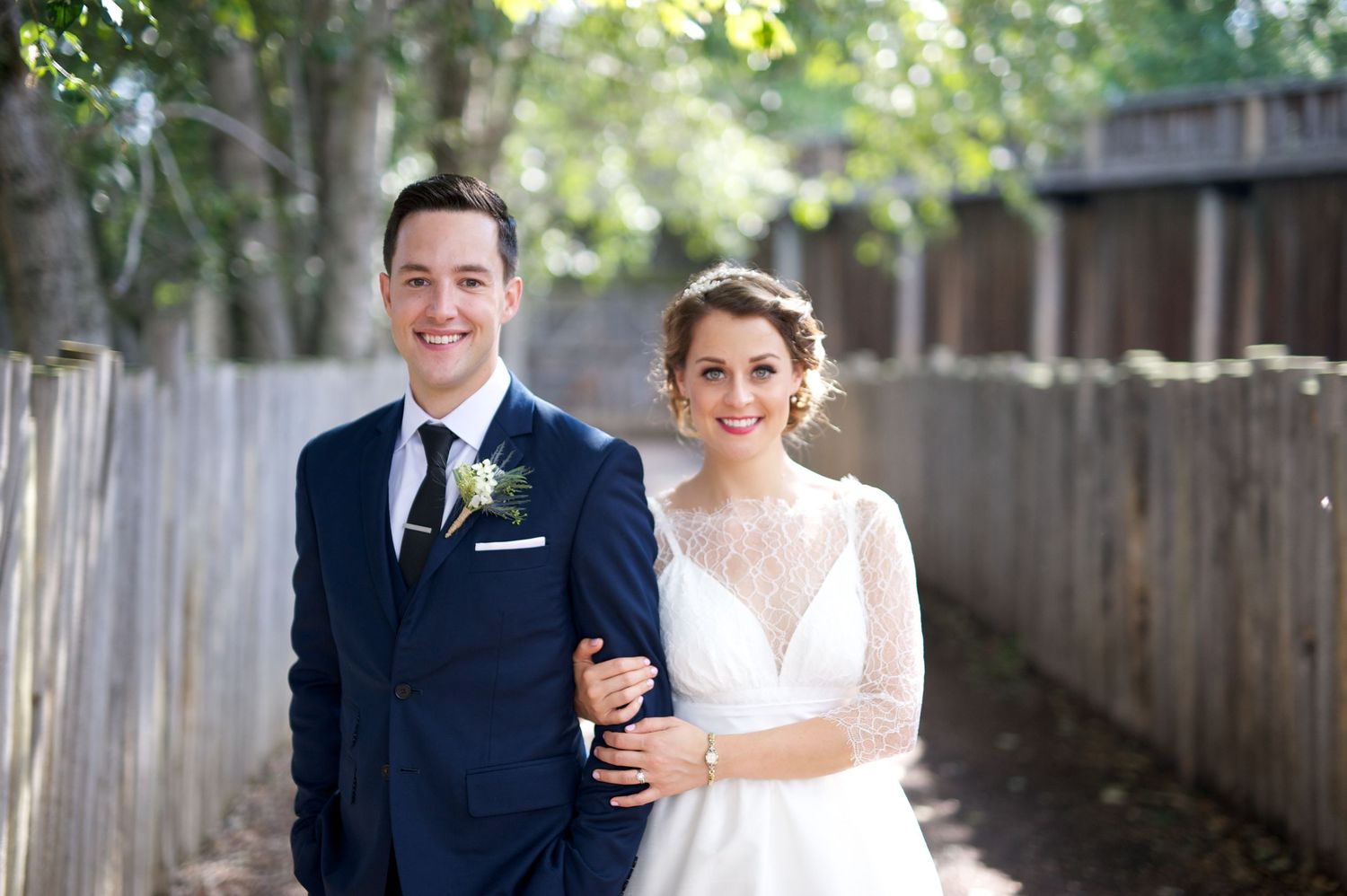 Bride and groom standing arm-in-arm on a tree-lined path between wooden fences, smiling warmly.