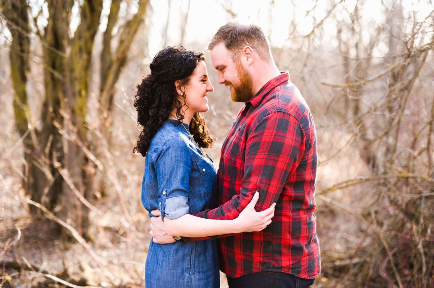 Couple standing face to face in early spring woods, wrapped in each other’s arms and sharing a soft, genuine smile.