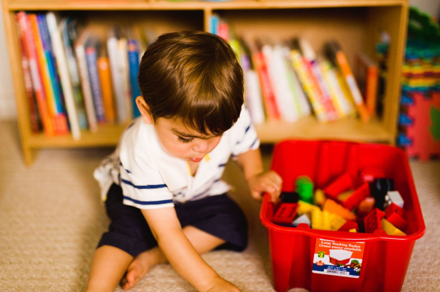 Young boy sitting on the floor, focused on playing with colorful toys from a red bin in front of a bookshelf.