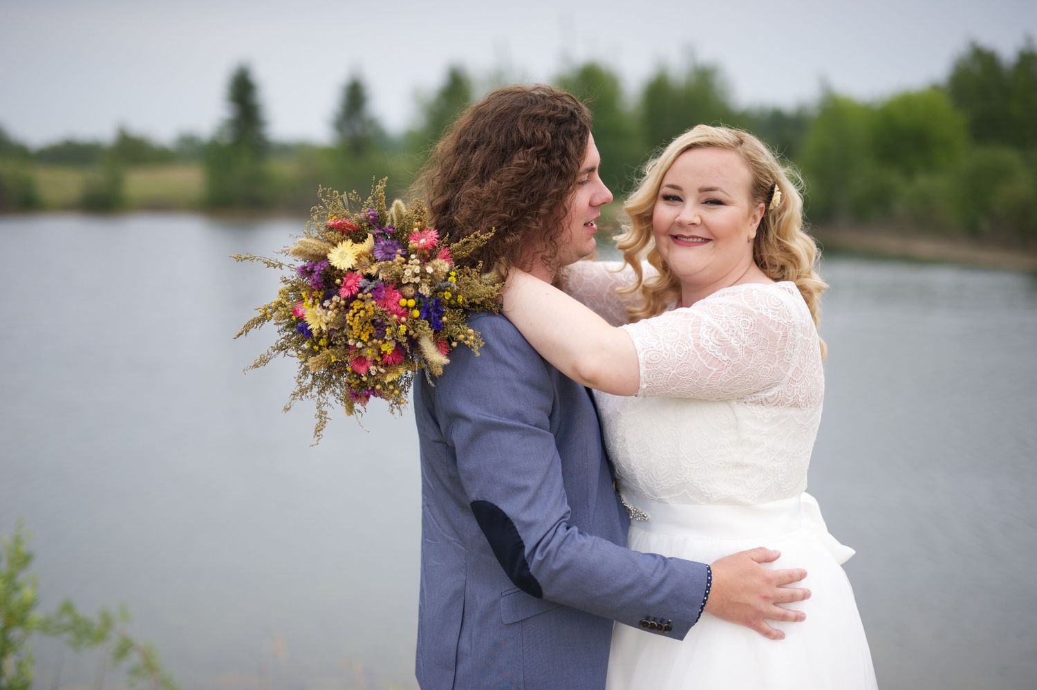 Bride hugging her groom beside a calm pond, bouquet draped over his shoulder, both looking serene and joyful.