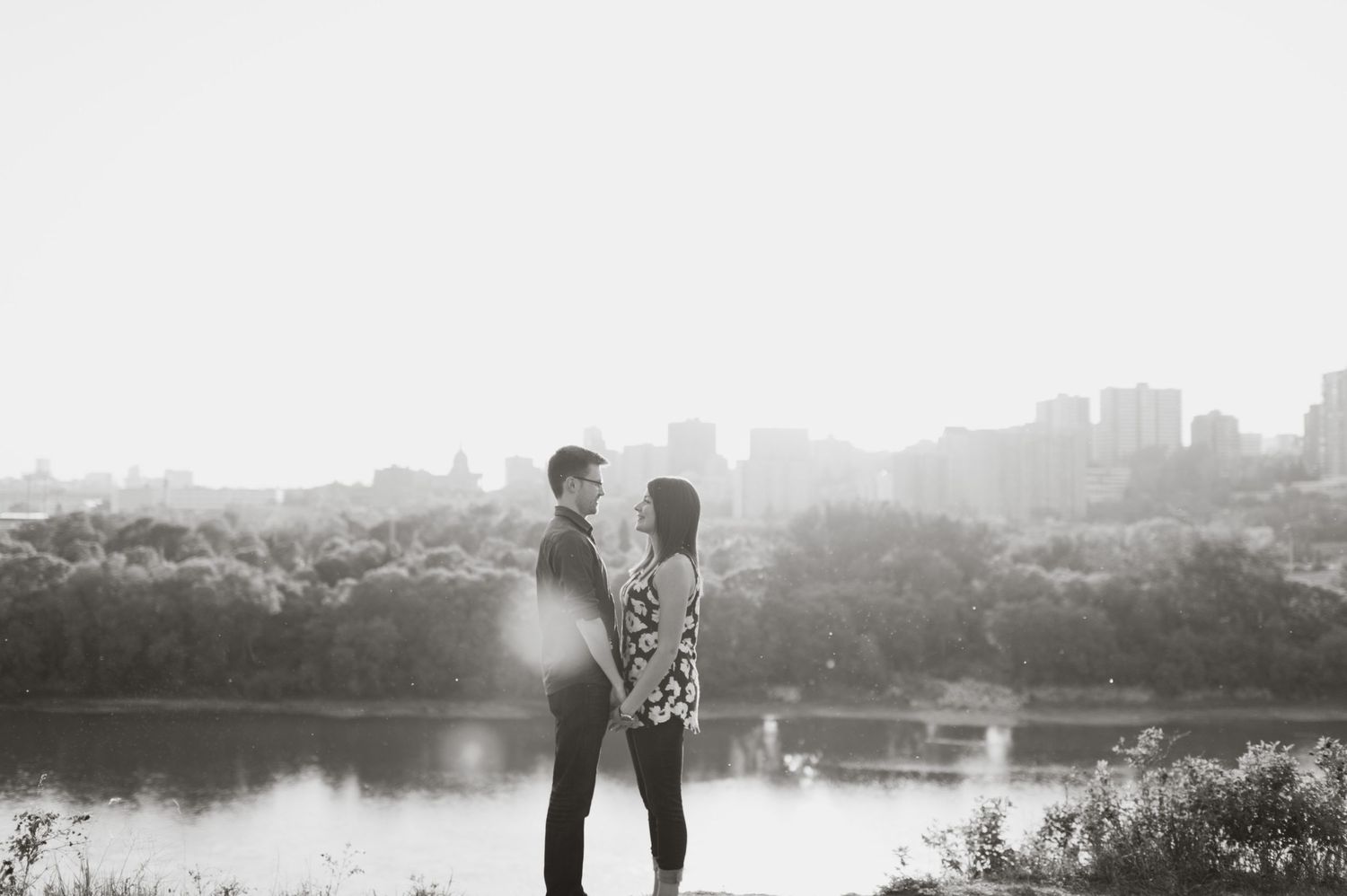 Black-and-white photo of a couple standing on a hilltop overlooking a river and distant city skyline.