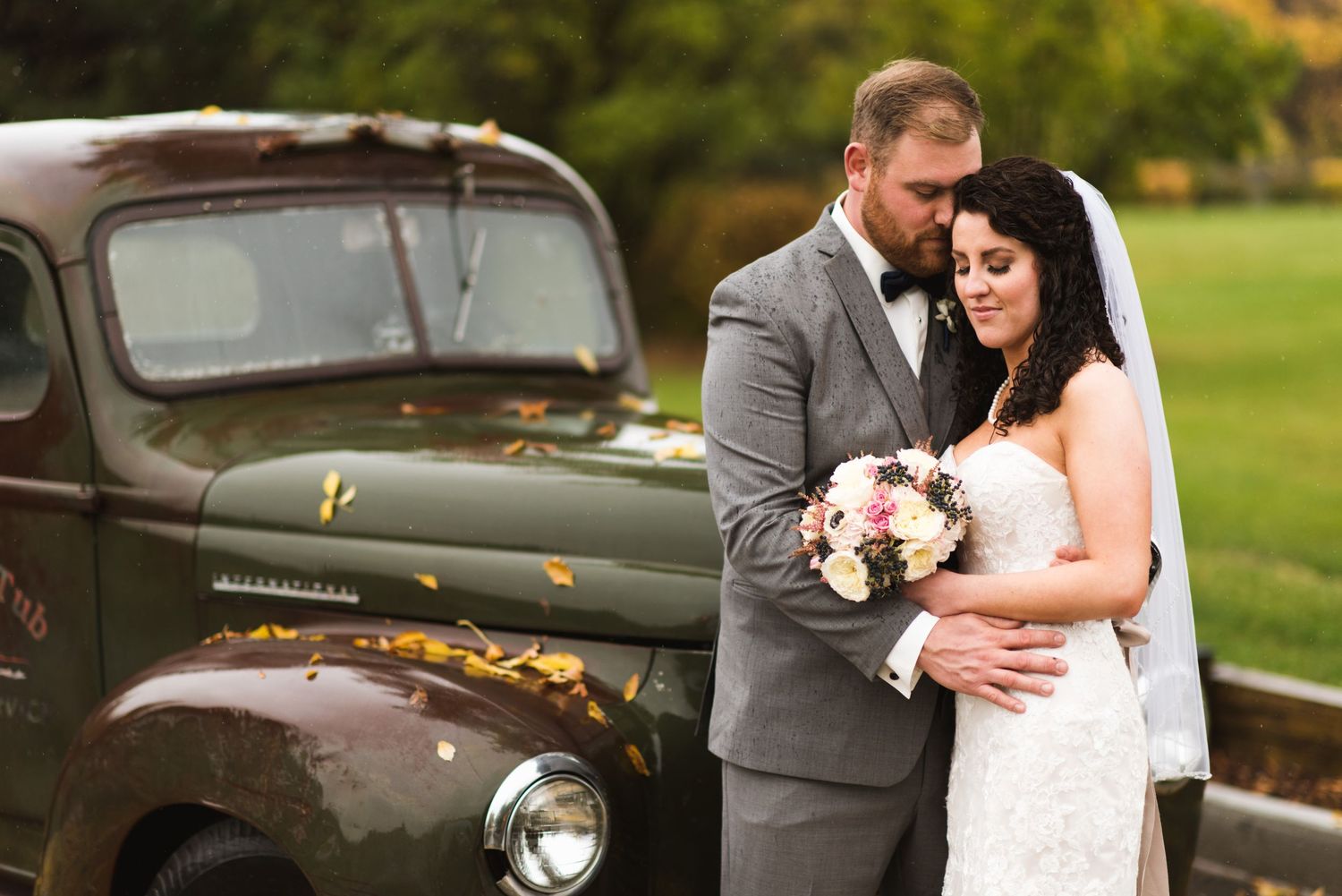 Bride and groom standing close together beside a weathered army-green vintage truck, leaves scattered on the hood, bouquet held between them