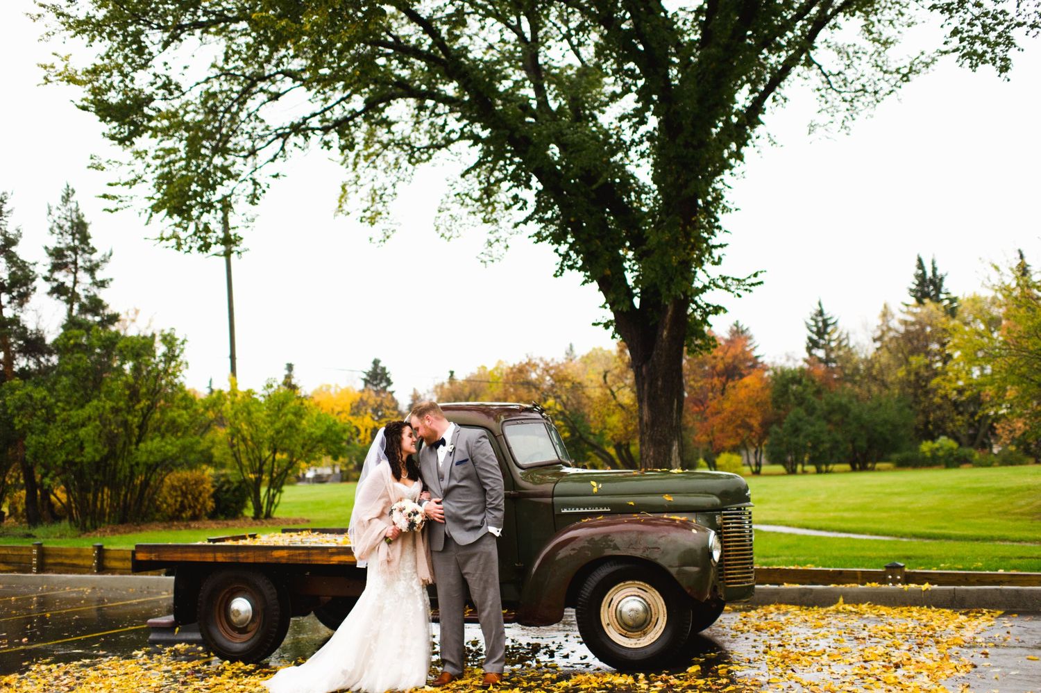 Bride and groom kiss beside an old pickup under a tree in a golden autumn park.