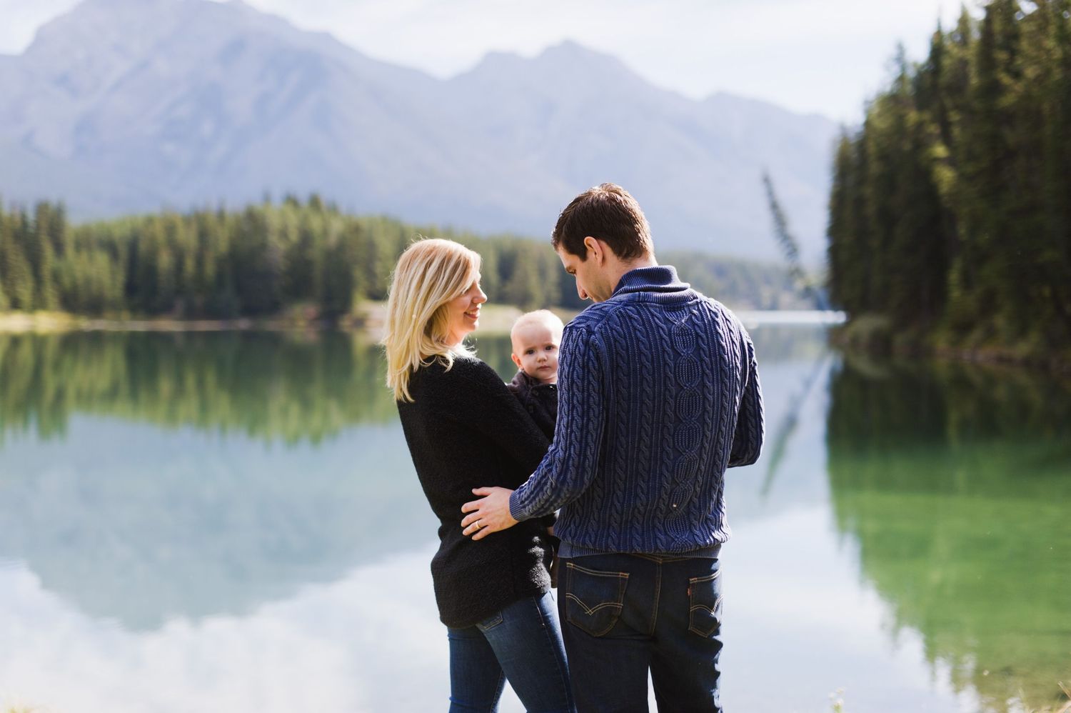 Young family poses beside reflective lake in the Rockies. Mountain family session with toddler in Alberta.