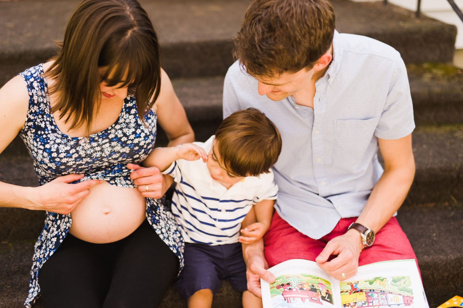 Little boy curiously touching mom's pregnant belly as parents sit with him reading on the steps.